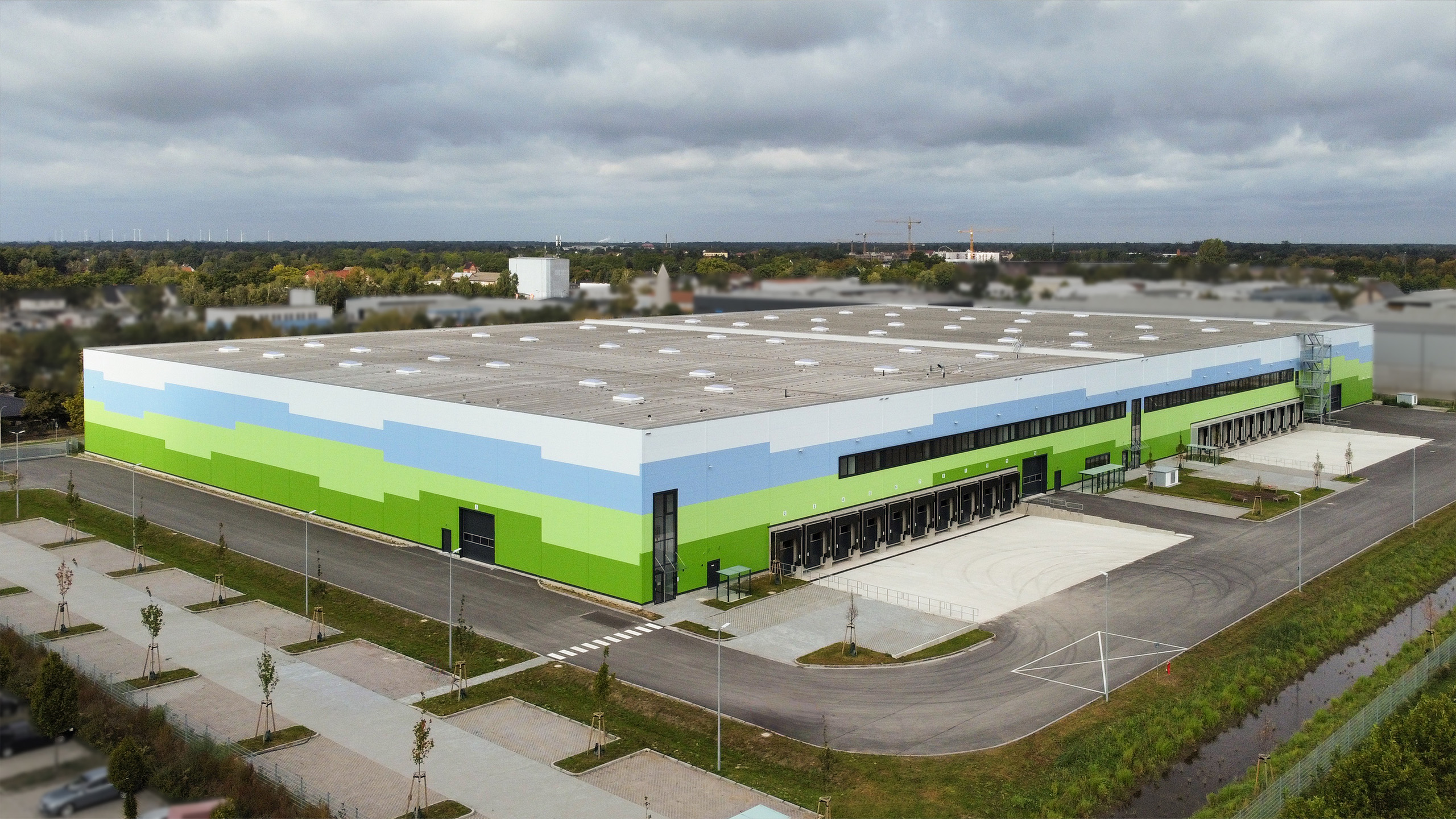 Modernes Lagergebäude mit buntem Fassadendesign in Grün- und Blautönen, umgeben von Parkplätzen und Vegetation.