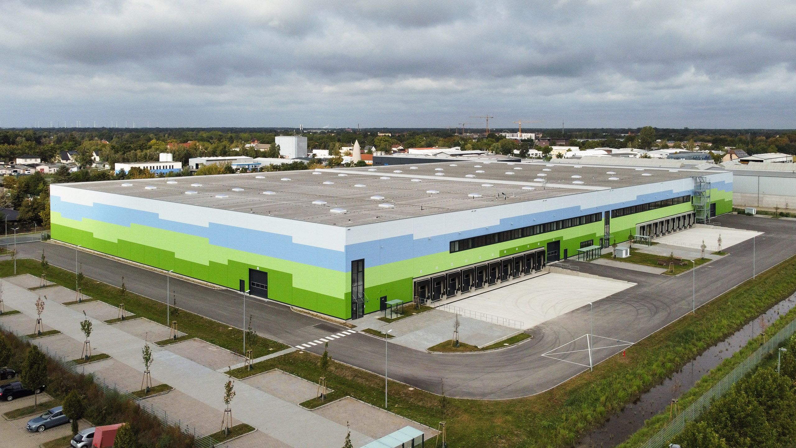 Farbenfrohes Logistikgebäude mit Ladebuchten, Parkplatz und umliegender Landschaft unter bewölktem Himmel.