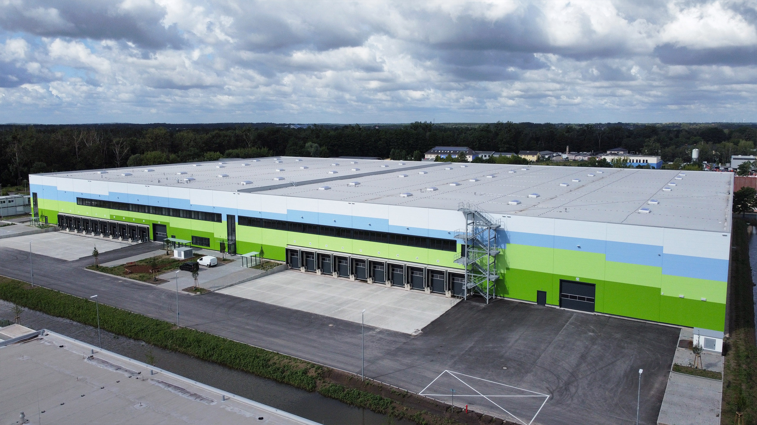 Luftaufnahme eines modernen Lagerhauses mit bunten Fassaden und Ladebereichen, umgeben von grüner Landschaft und Wolken am Himmel.