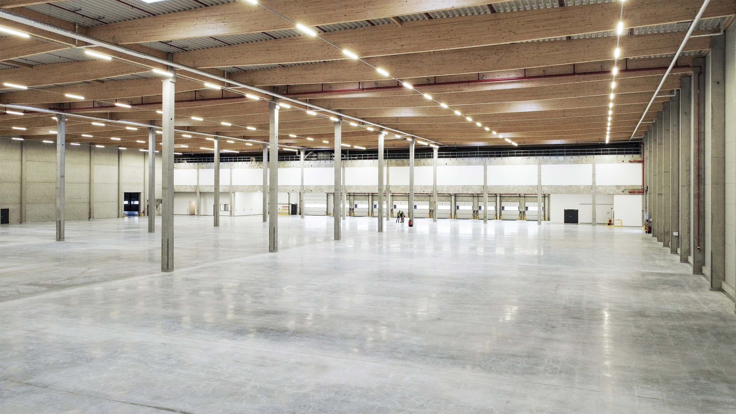 Großer, leerer Industrie- oder Lagerraum mit Betonboden und Holzdecke, beleuchtet von mehreren Deckenlampen.
