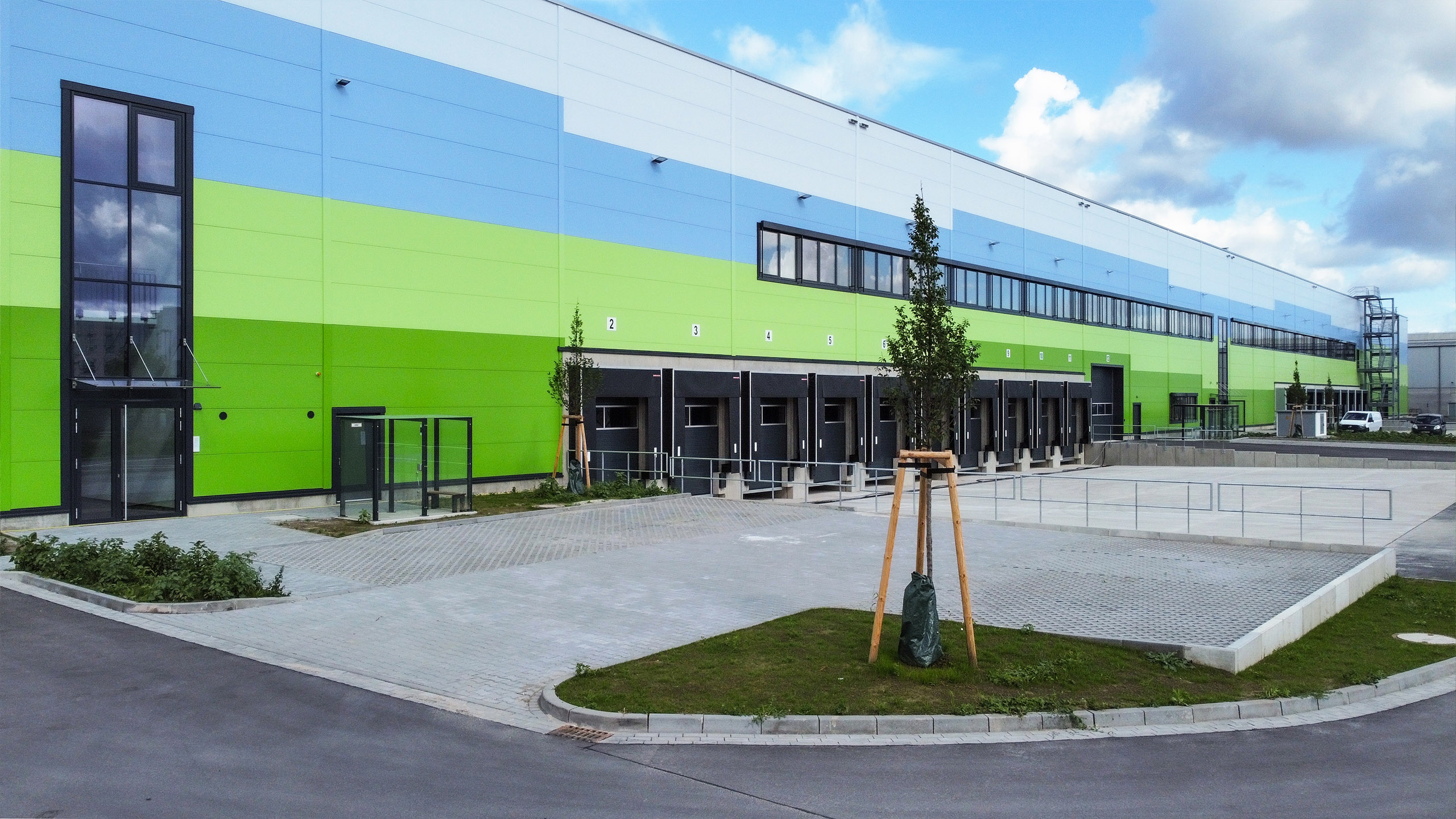 Neues, modernes Lagerhaus mit grün-blauer Fassade, Ladebereichen und begrüntem Außenbereich.