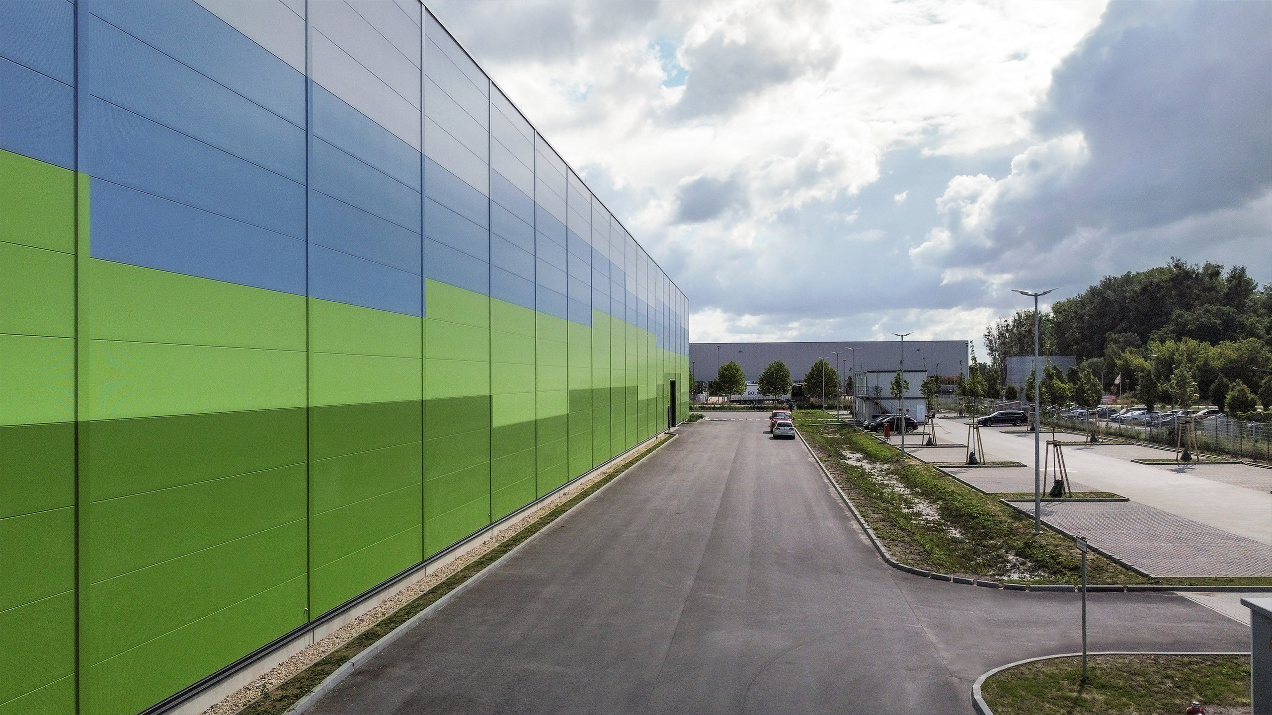 Farbige Industriegebäudewand mit grünen und blauen Elementen, daneben asphaltierte Straße und parkende Autos.