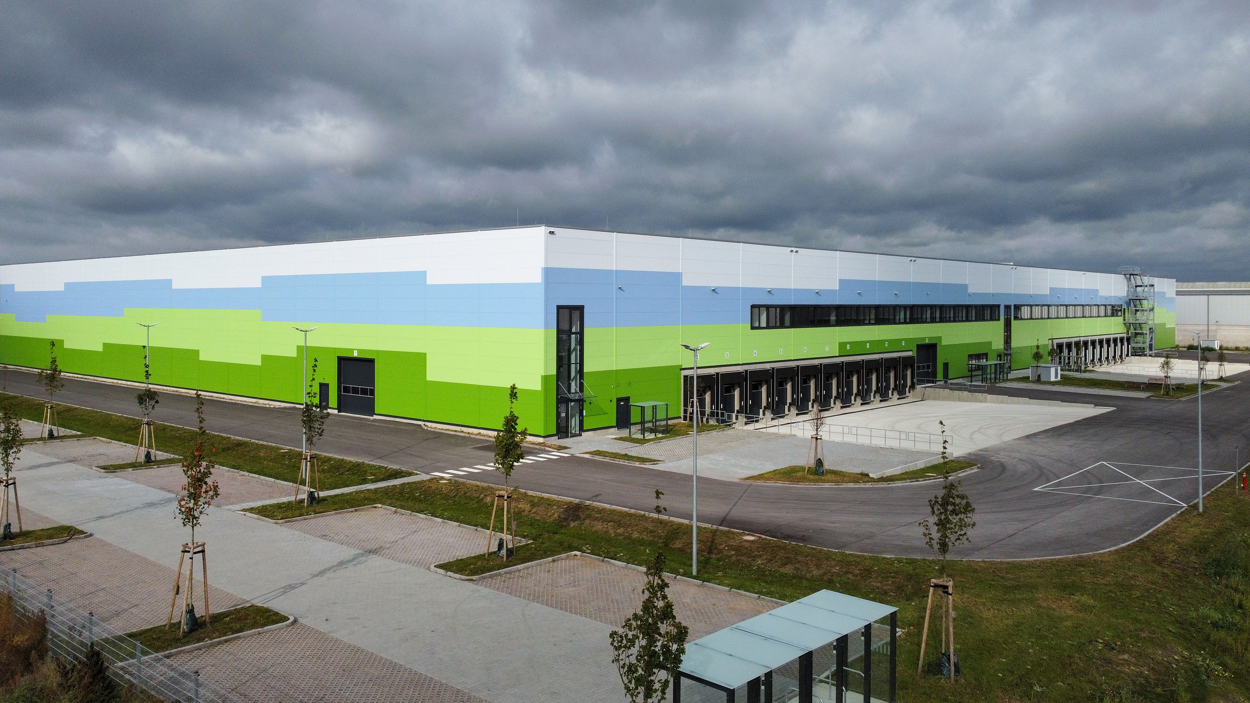 Große moderne Lagerhalle mit buntem Fassadendesign in Grün- und Blautönen, umgeben von kleinen Bäumen und Parkplatzflächen.
