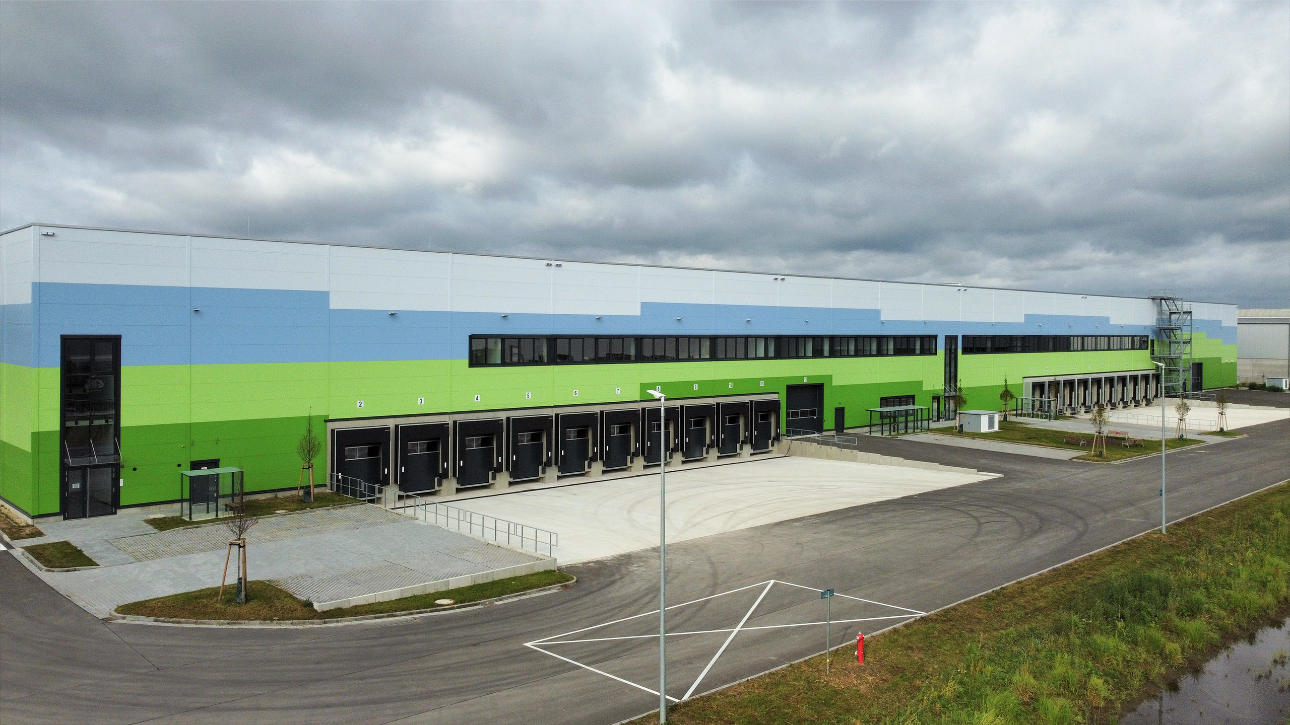 Großes, modernes Lagergebäude mit farbigen Wänden, Laderampen und gepflastertem Vorplatz unter bewölktem Himmel.