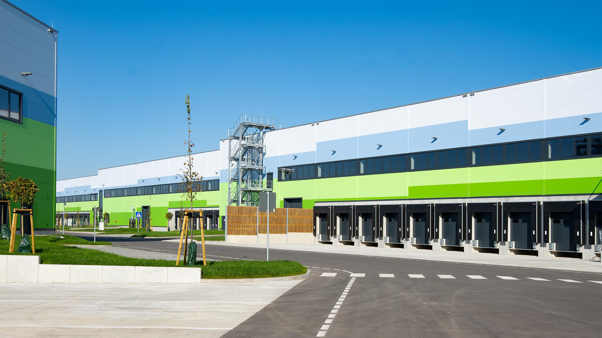 Moderne Lagergebäude mit grüner und blauer Fassade, angrenzende Ladebereiche und klar blauer Himmel.