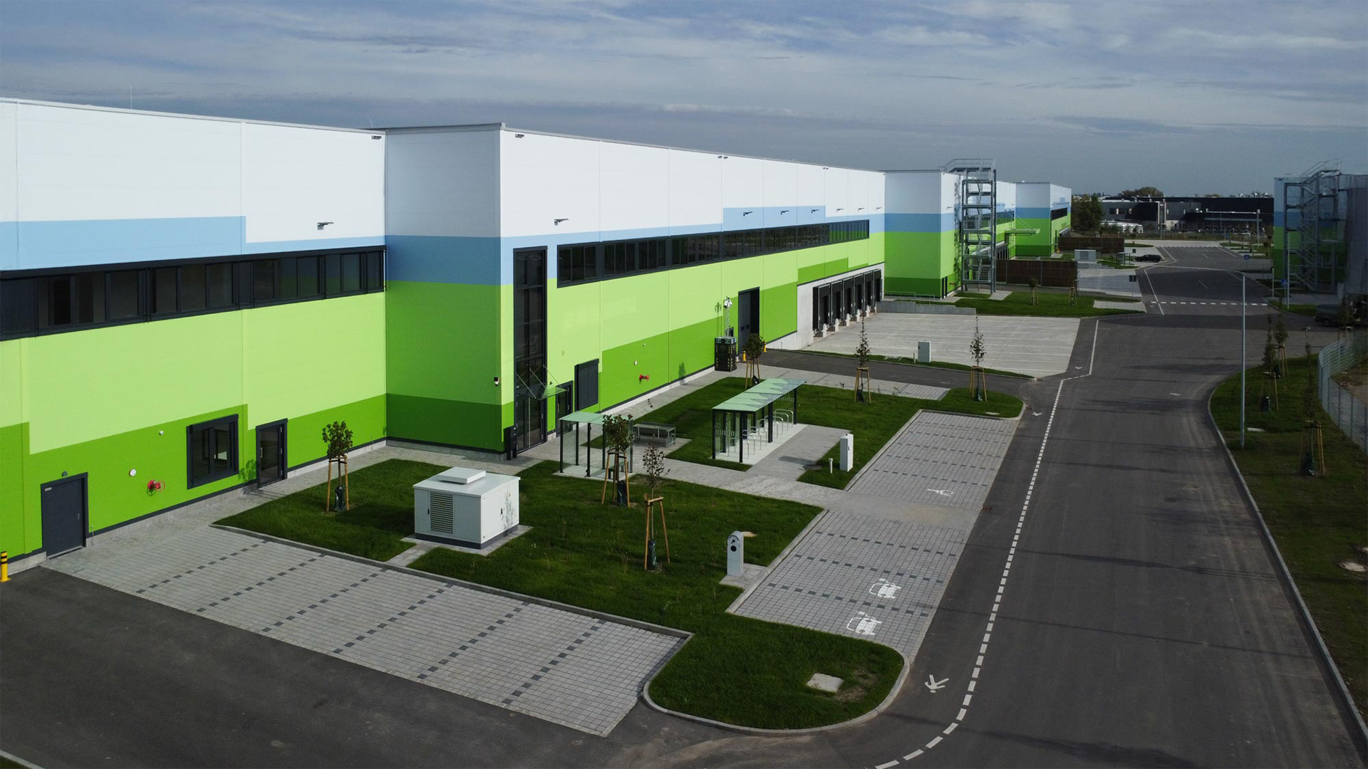 Moderne Lagerhalle mit grün-weißer Fassade, Zufahrtsstraße und begrüntem Bereich im Vordergrund.