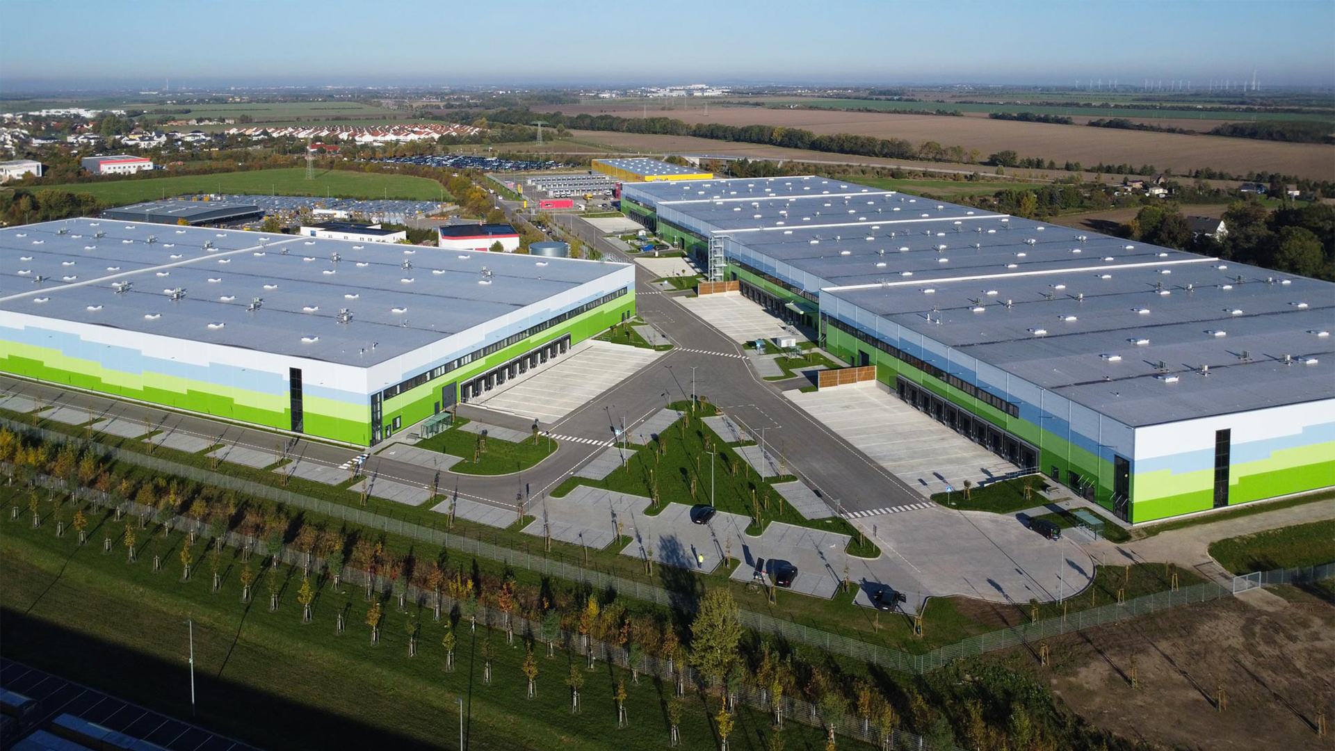 Luftaufnahme eines modernen Gewerbegebiets mit mehreren Lagerhallen und befestigten Parkplätzen, umgeben von Grünflächen.