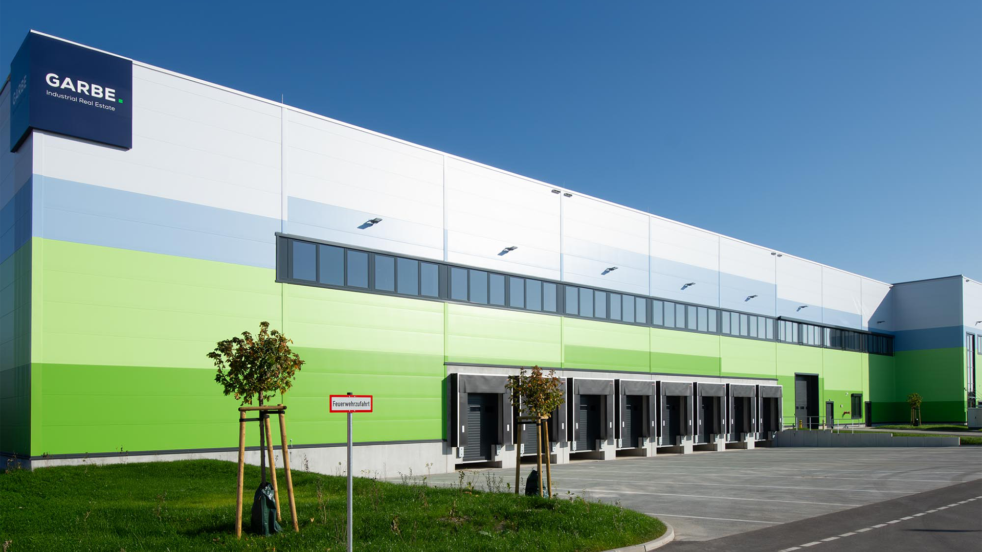 Moderne Lagerhalle mit grün-blauer Fassade, Fensterfront und Laderampen, umgeben von Grünflächen und Bäumen.