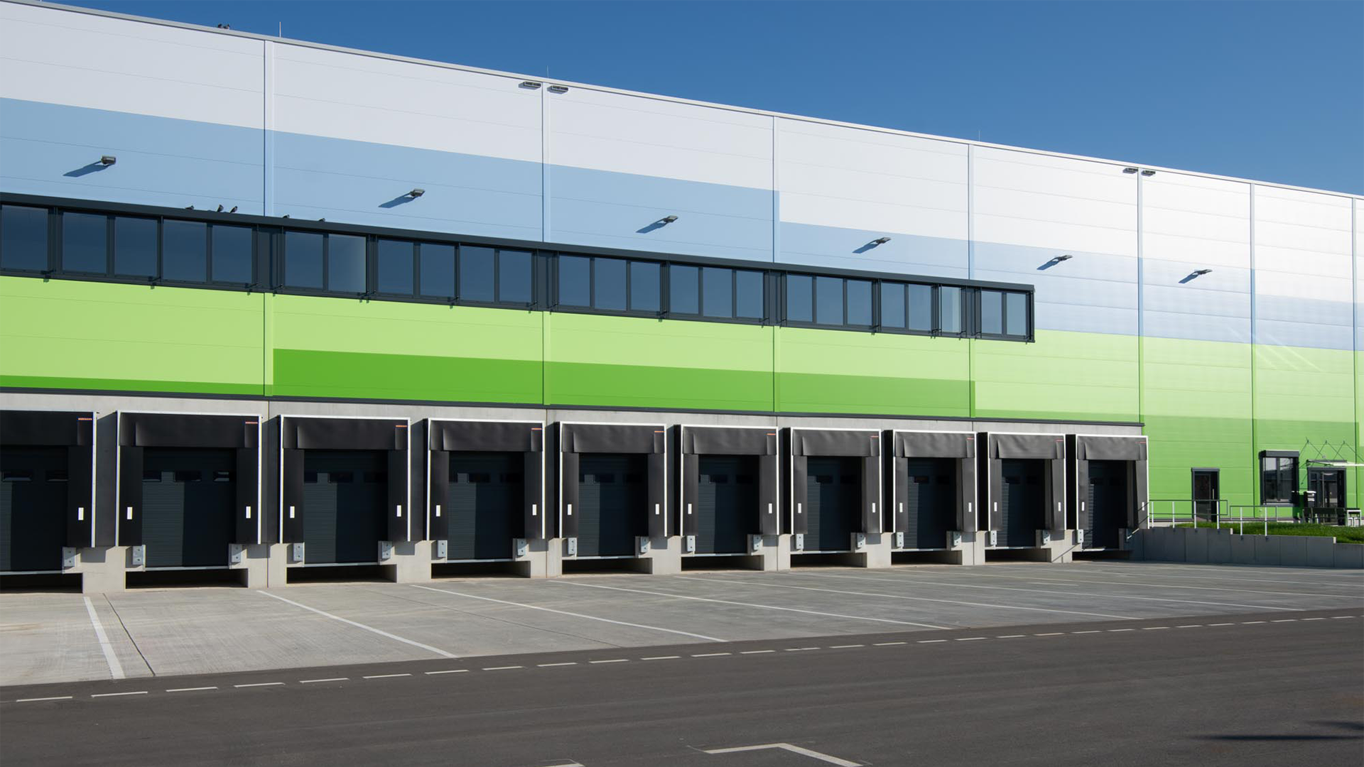 Moderne Logistikhalle mit grüner und blauer Fassade, mehreren Ladebuchten und klarem blauen Himmel.