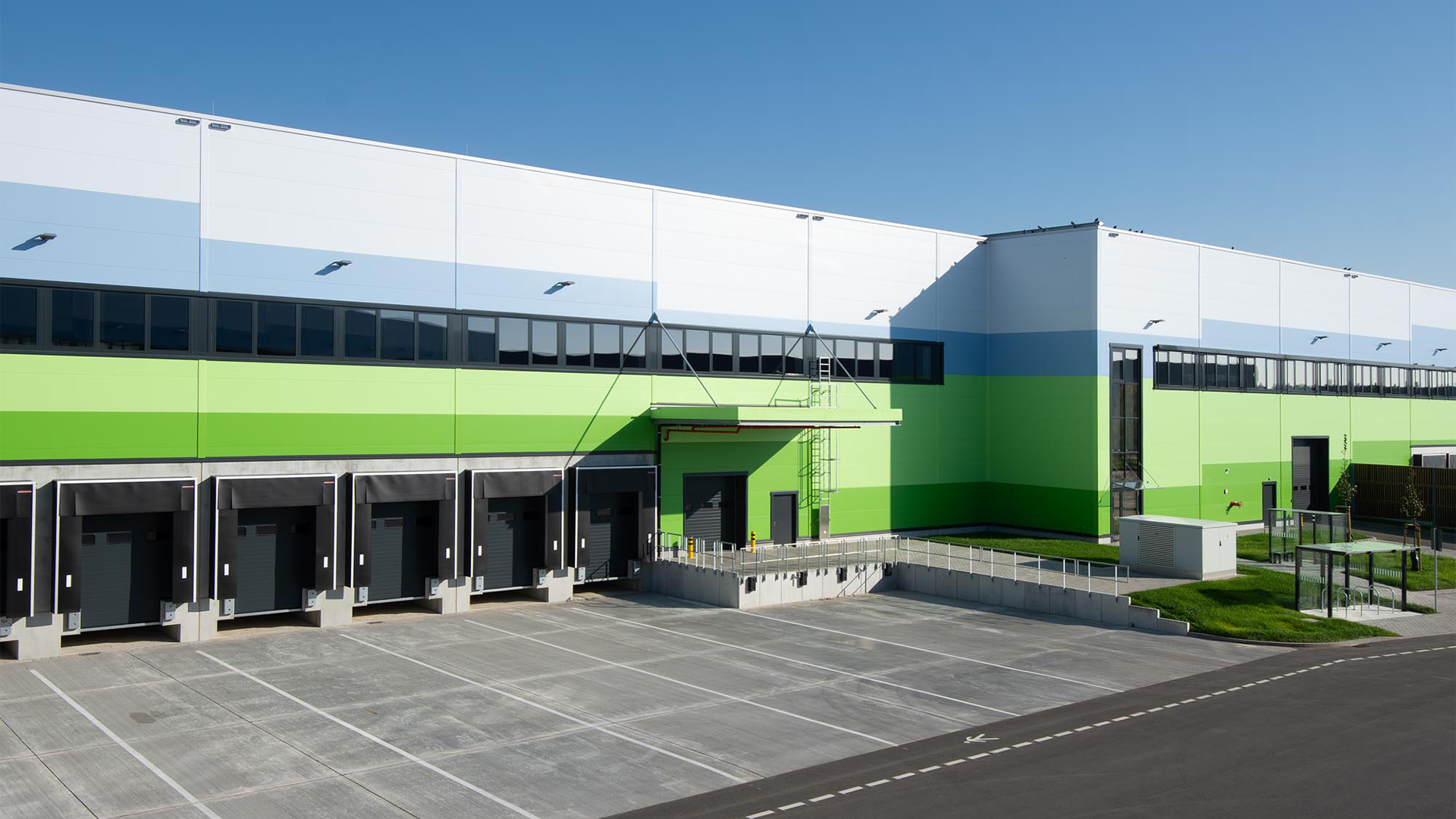 Moderne Lagerhalle mit grün-blauer Fassade und Verladerampen, umgeben von einem Parkplatz und Grünflächen.