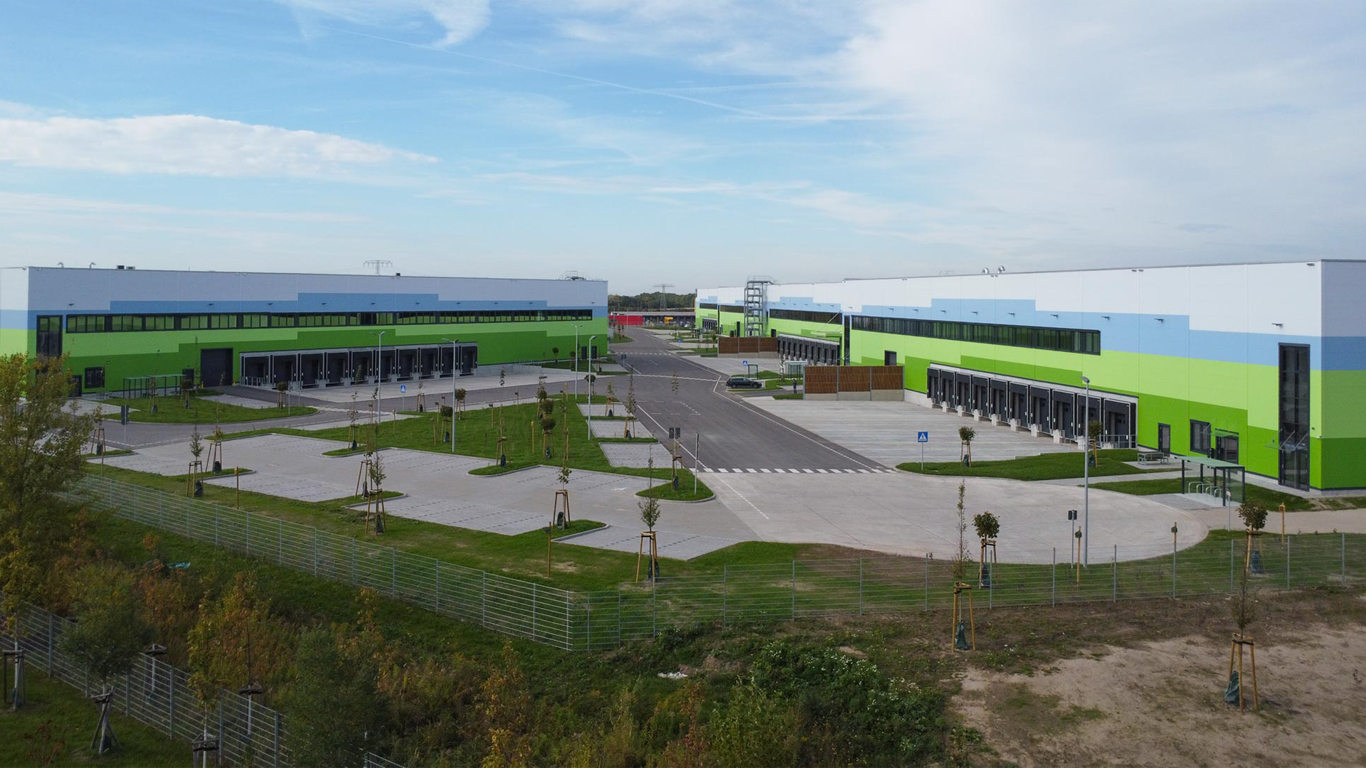 Luftaufnahme eines modernen Logistikgebäudes mit grüner und blauer Fassade, umgeben von Parkplätzen und Bäumen.