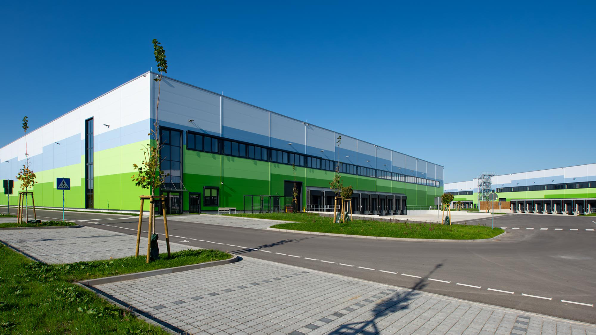 Moderne Lagerhalle mit grün-blauer Fassade und Bäumen im Vordergrund, unter strahlend blauem Himmel.