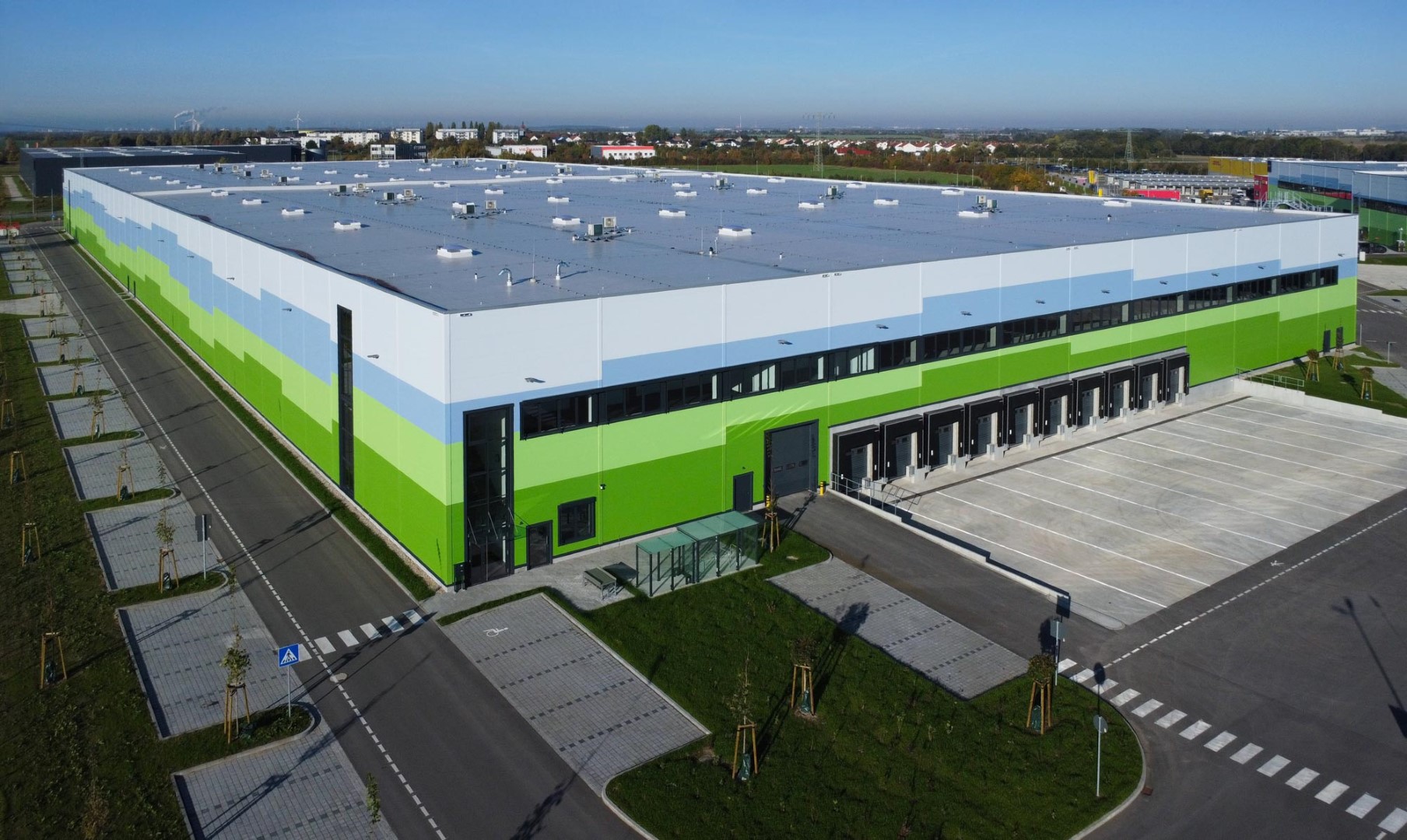 Moderne Lagerhalle mit farbigen Fassaden in Grün und Blau, umgeben von Parkplätzen und einer Zufahrt.