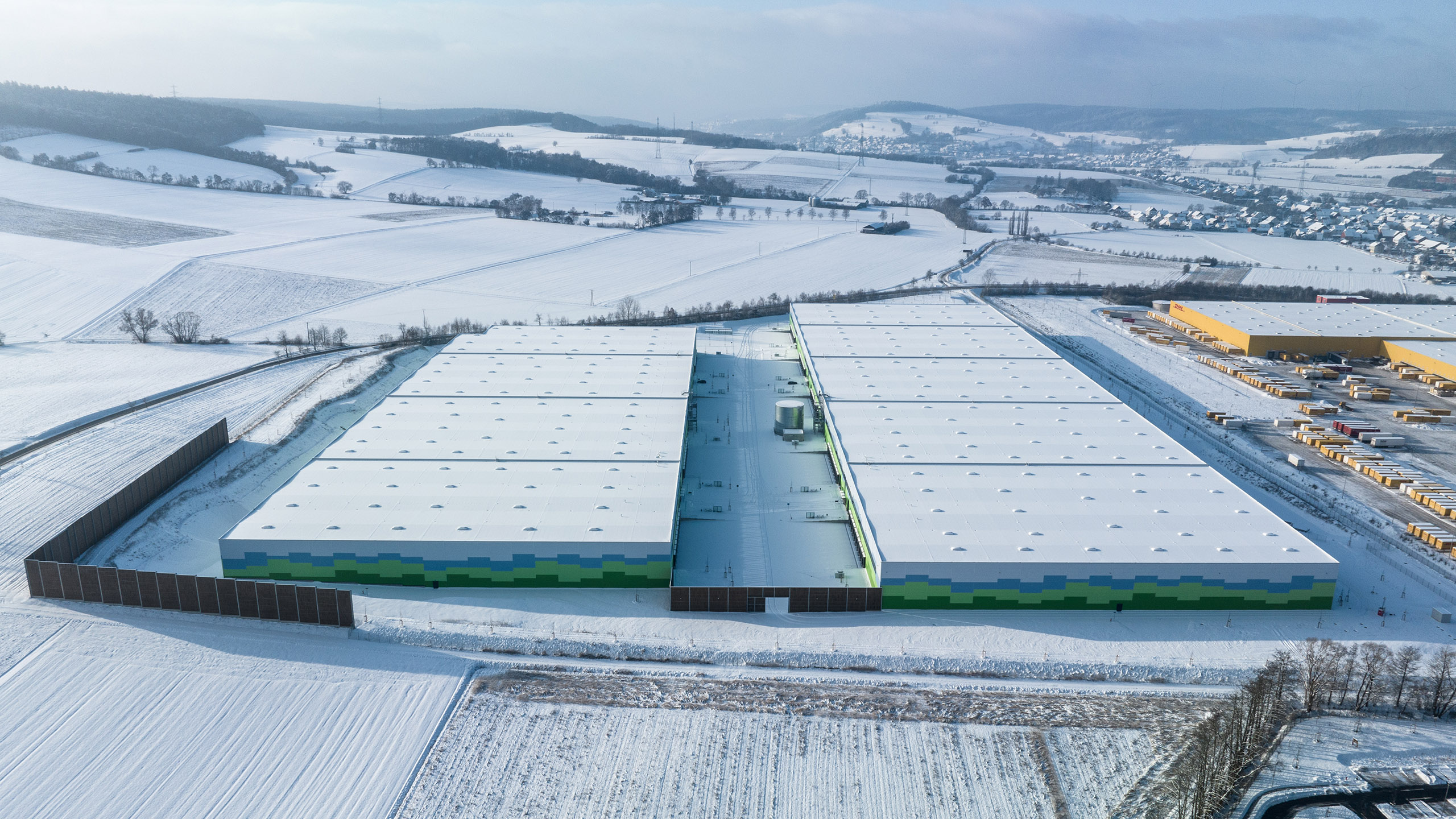 Luftaufnahme eines großen Lagerhauses mit grün-blau gestreiften Wänden, umgeben von verschneiten Feldern und Hügeln.