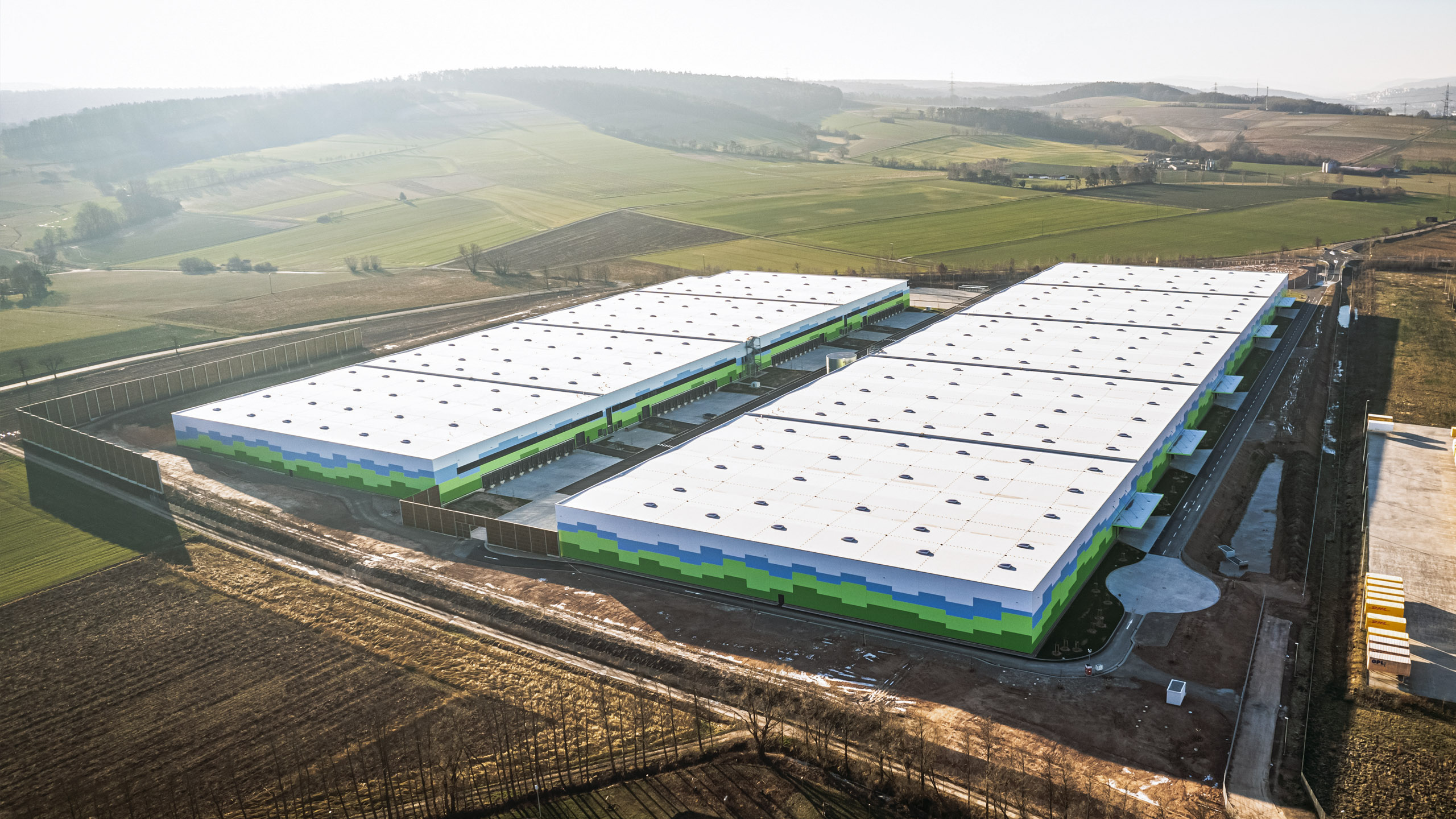 Luftaufnahme eines großen Lagergebäudes mit bunten Fassaden, umgeben von grünen Feldern und Hügeln.
