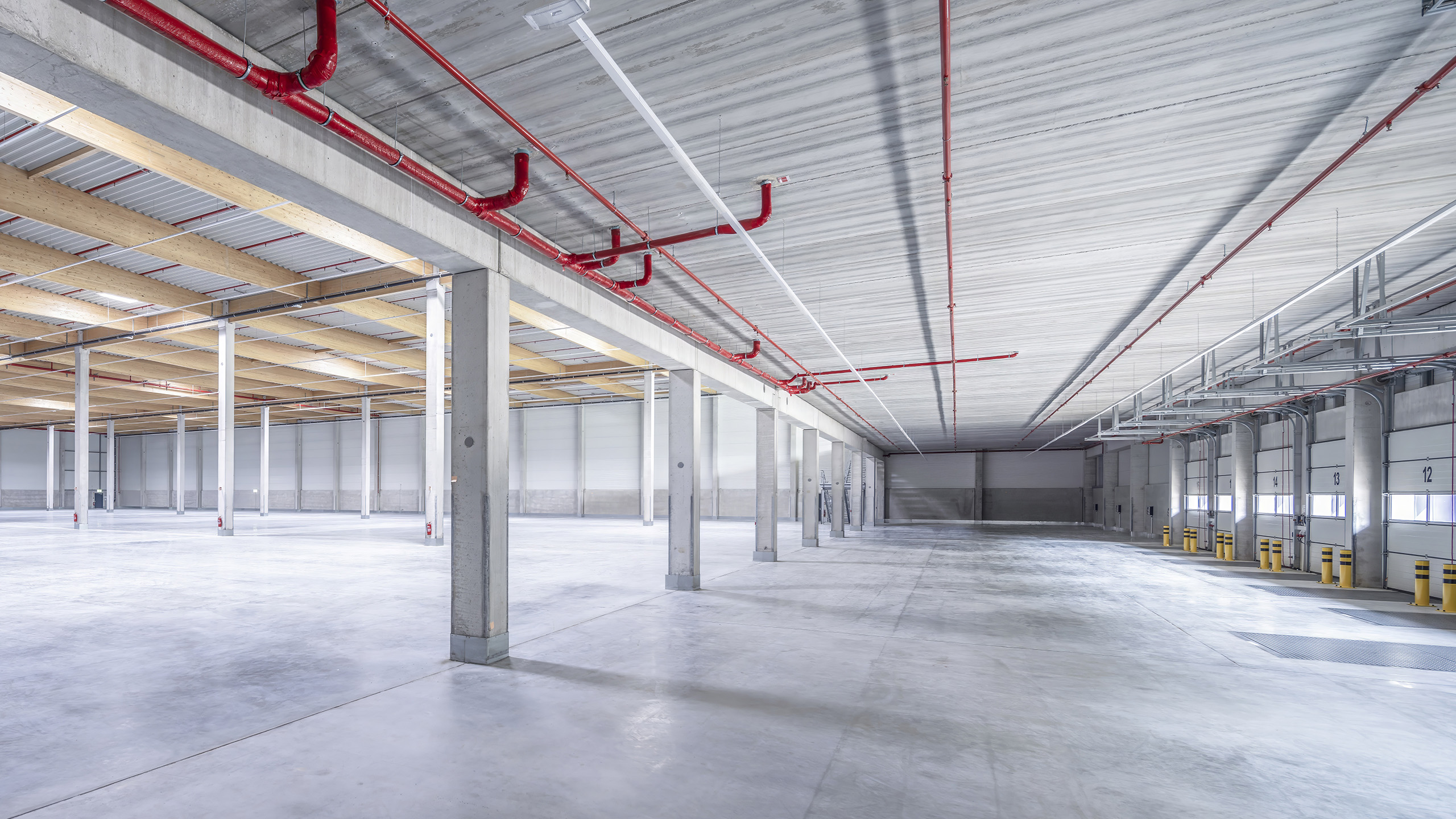 Geräumiger Lagerraum mit Betonboden, Stahlstützen und roten Rohrleitungen an der Decke, beleuchtet durch Oberlichter.
