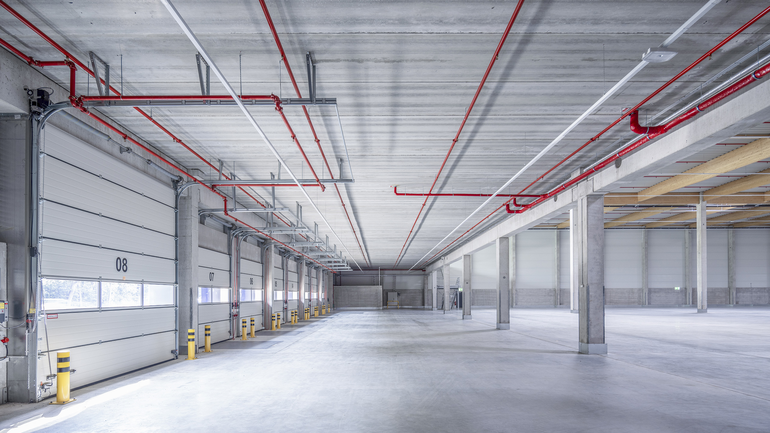 Großer, leerer Lagerraum mit grauem Betonboden, großen Türen und roten Rohren an der Decke.