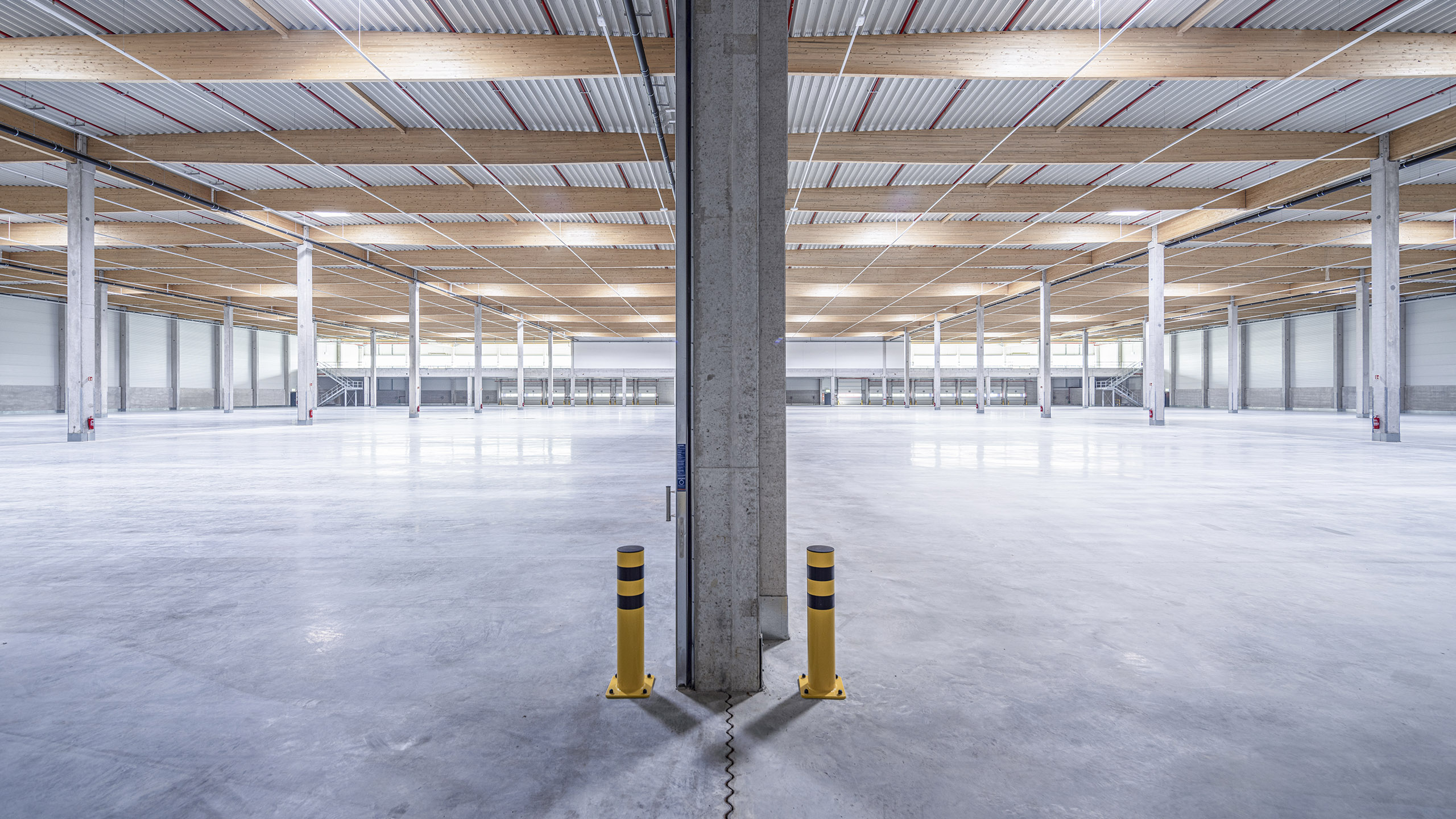 Leerer Lagerraum mit hohen Decken, Holz- und Betonstrukturen sowie gelben Pollern im Sichtfeld.