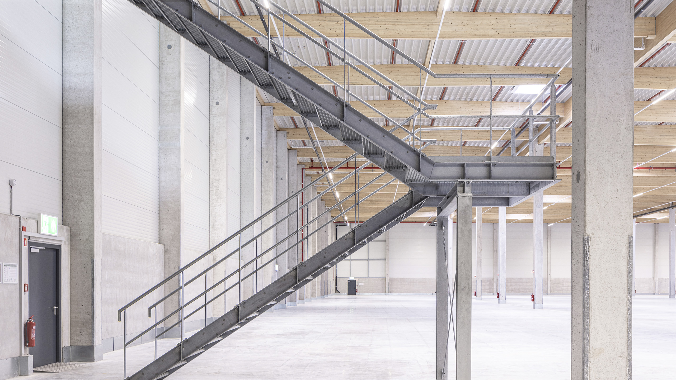 Stahltreppe in einem modernen Lager mit Betonwänden und Holzdecke, gut beleuchtet und mit Notausgang.