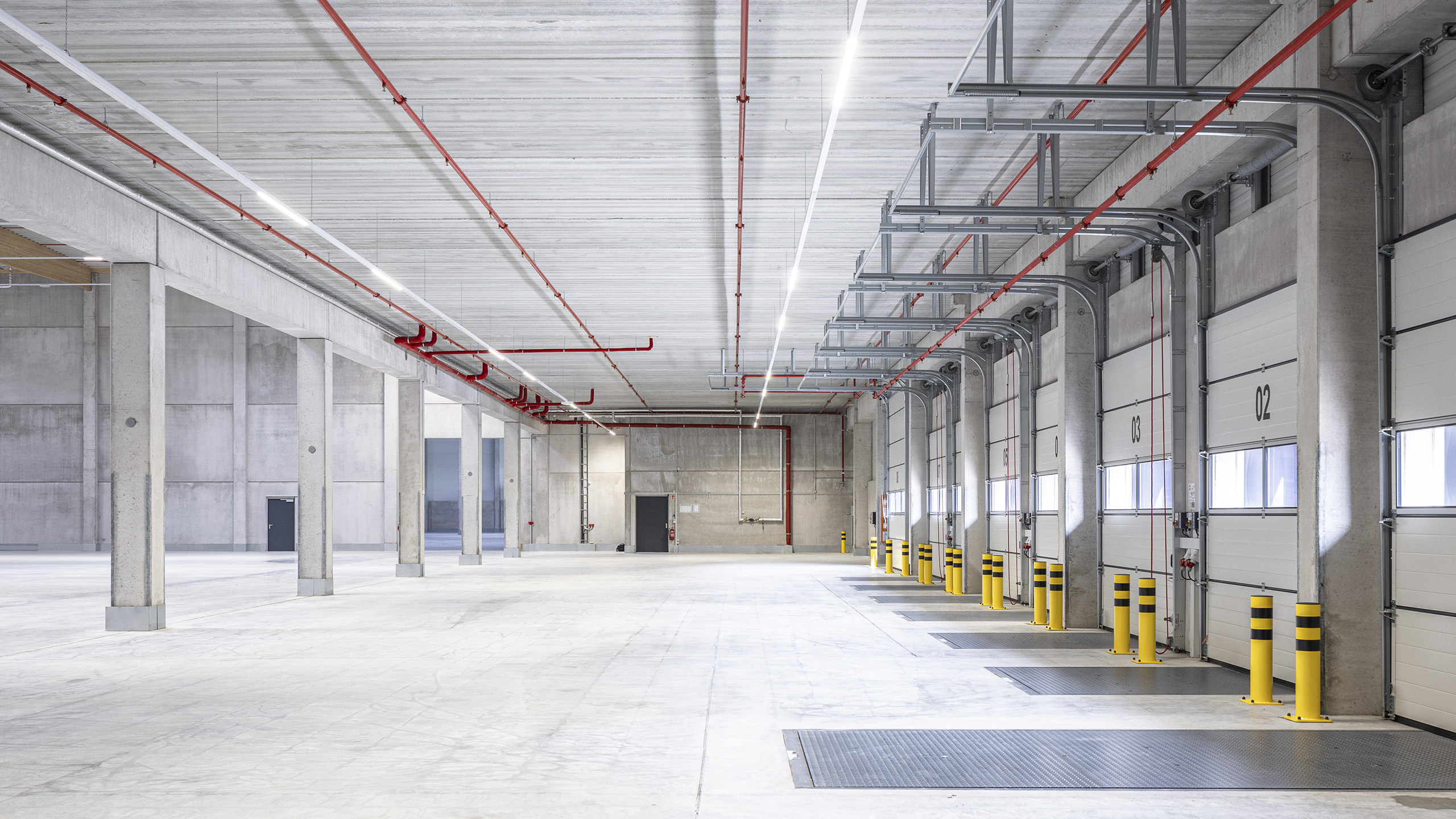 Großer, heller Lagerraum mit grauen Wänden, gelben Pollern und sichtbaren Rohren an der Decke.