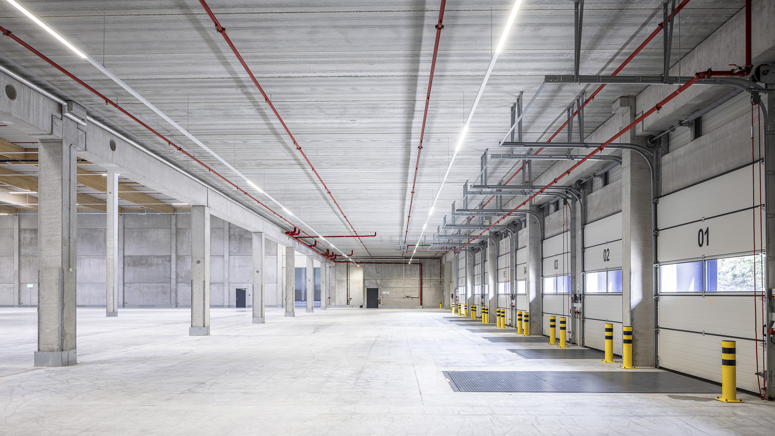 Heller, moderner Lagerraum mit Betonwänden, hohen Decken und mehreren Rolltoren, gekennzeichnet durch gelbe Sicherheitsständer.
