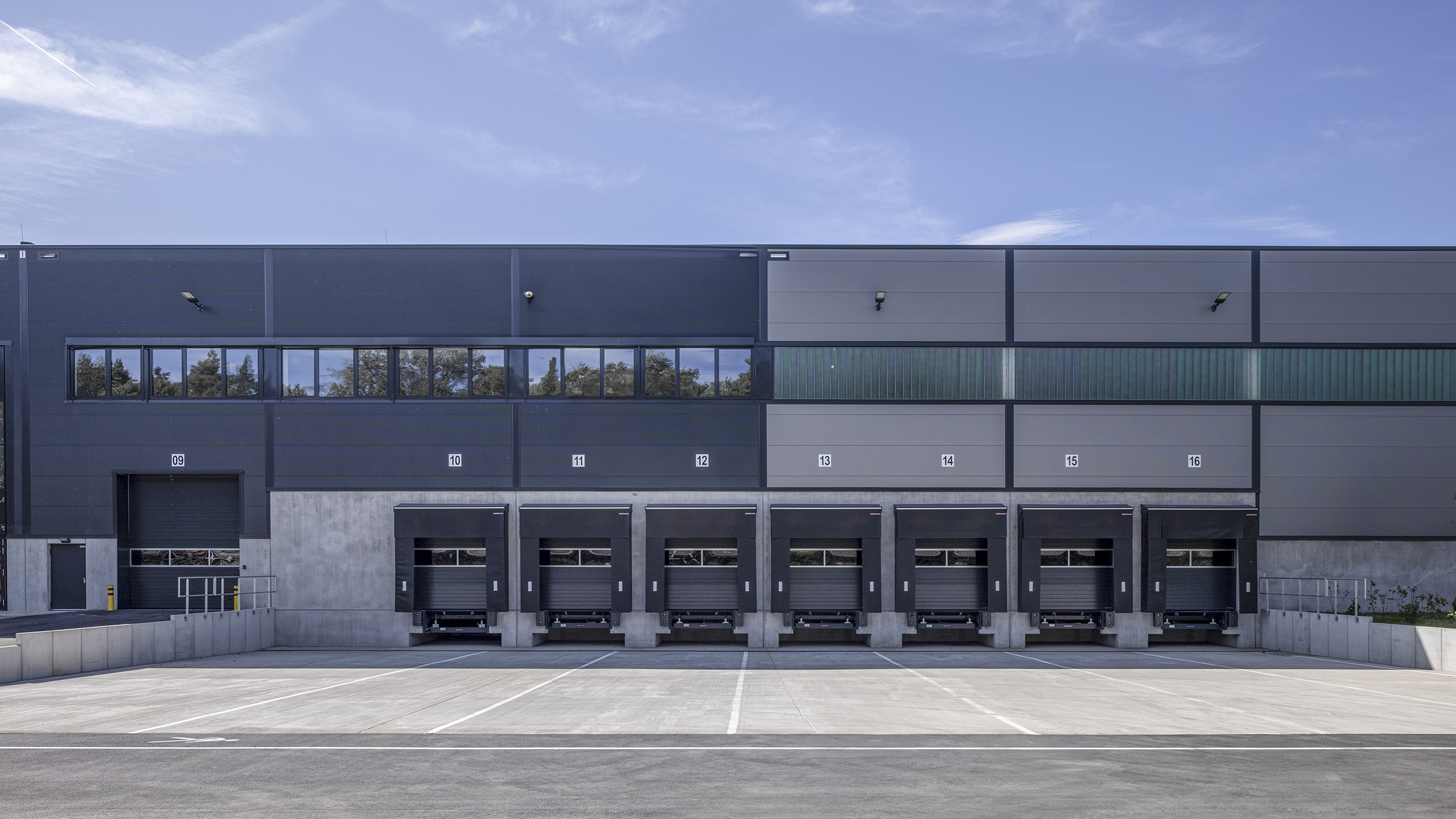 Moderne Lagerhalle mit Laderampen, klarer Himmel, graue Fassade und Parkplatz im Vordergrund.