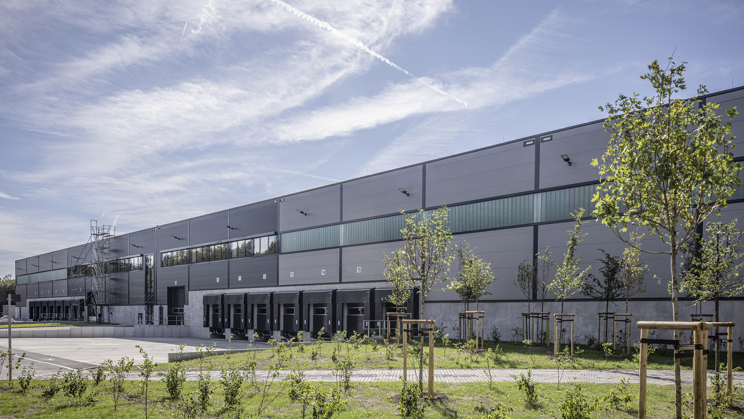 Moderne Lagerhalle mit großen Fenstern, umgeben von Grünflächen und kleinen Bäumen unter blauem Himmel.