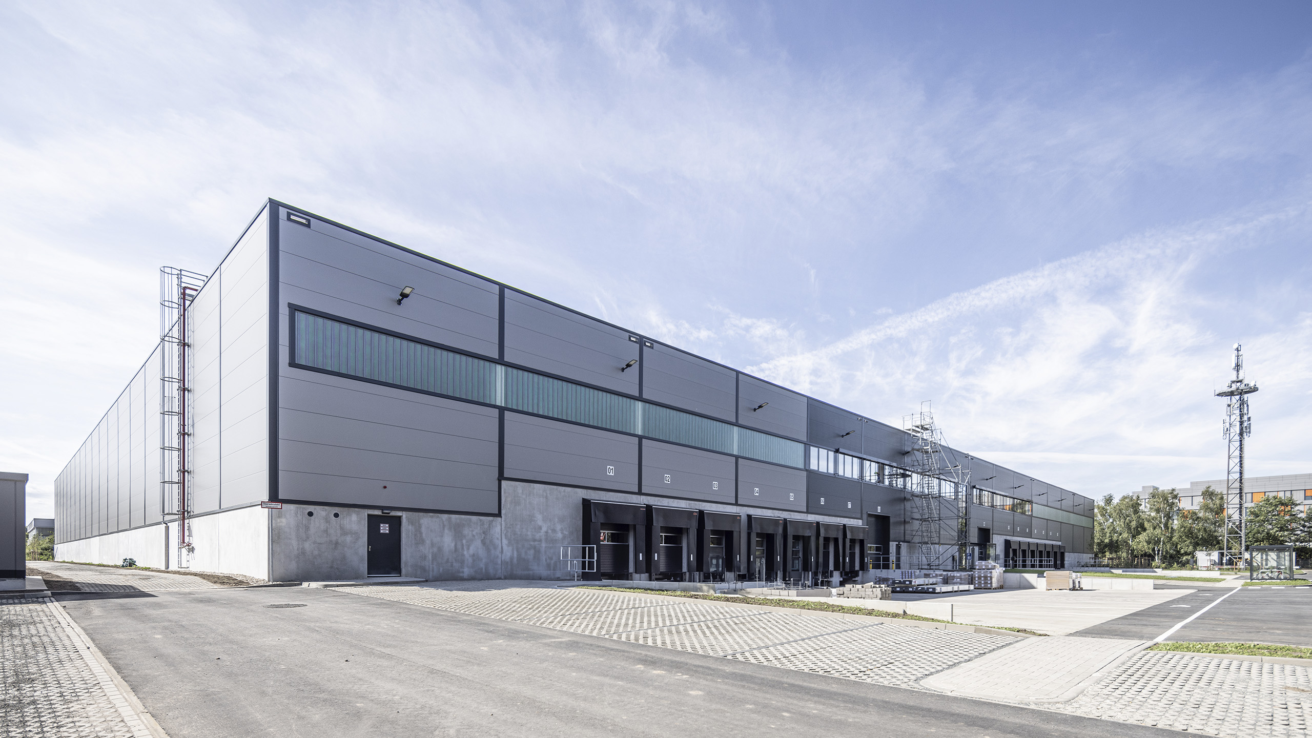 Modernes Lagergebäude mit Fenstern und Rampe, umgeben von gepflastertem Gelände und Hochspannungsmasten.