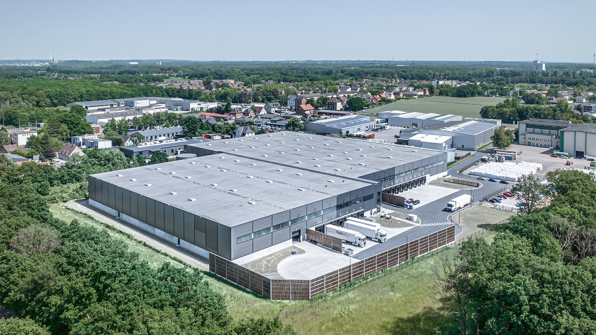 Luftaufnahme eines modernen Lagerhauses mit umliegenden Gebäuden und Bäumen in ländlicher Umgebung.