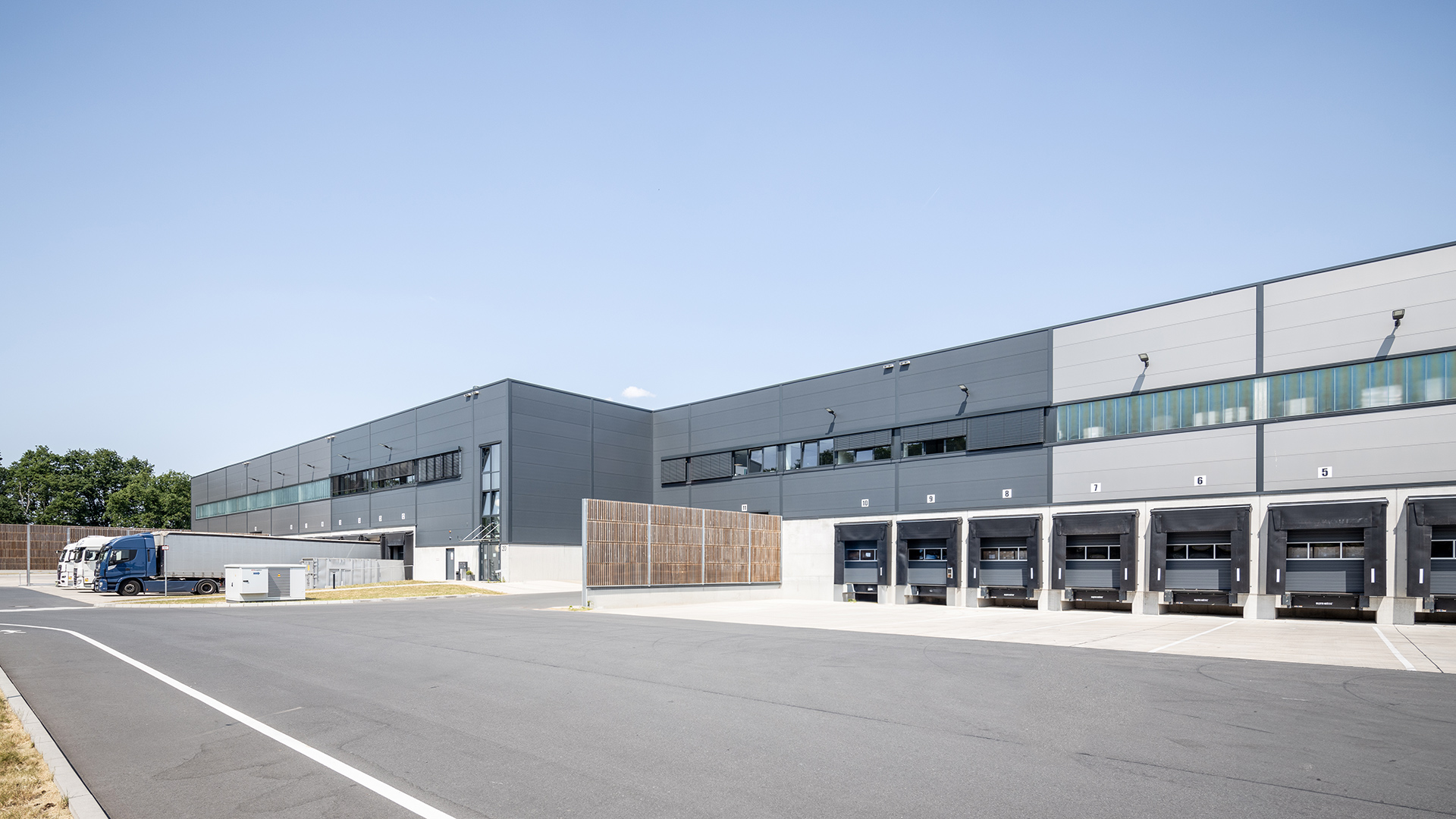 Moderne Lagerhalle mit Lkw-Entladezonen und großen Fenstern unter blauem Himmel.