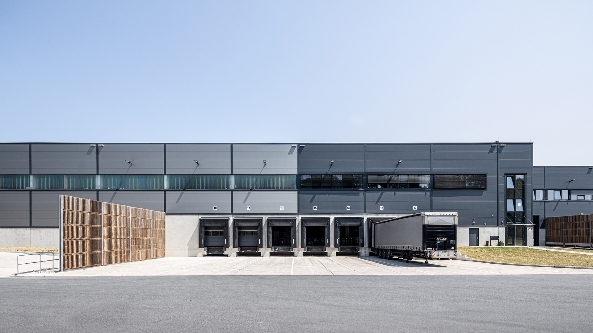 Lagergebäude mit mehreren Laderampen und einem Lastwagen auf dem Hof, umgeben von einem Holzzaun und klarem Himmel.