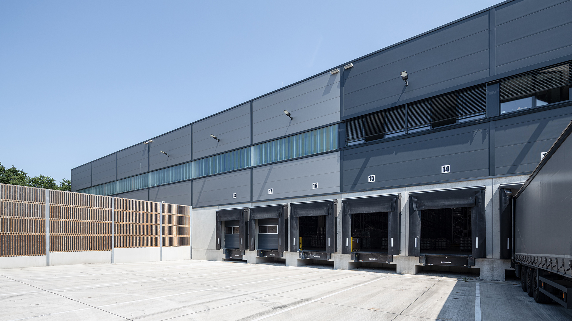 Moderne Lagerhalle mit Ladebuchten, grauer Fassade und klar blauem Himmel, teilweise mit Lkw beladen.