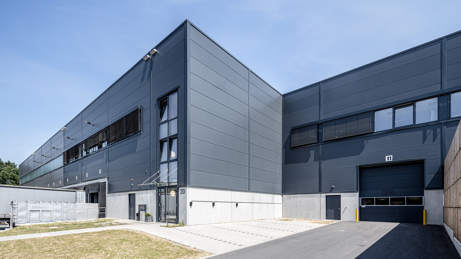 Moderne Lagerhalle mit grauer Fassade, großen Fenstern und einem Ladedock. Grundstück mit Rasenfläche und gepflastertem Zugang.
