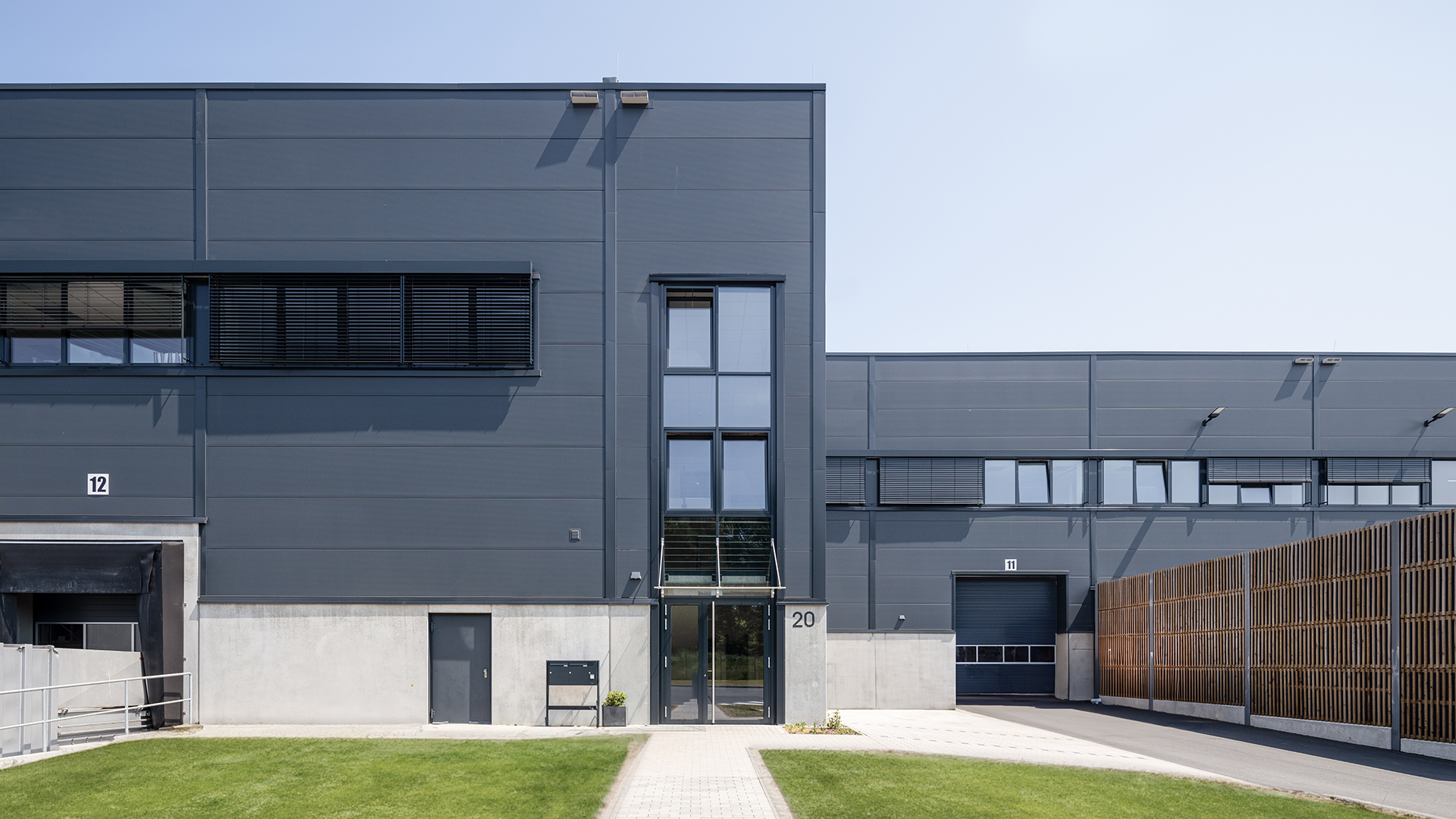 Modernes Industriegebäude mit glatten grauen Wänden, großen Fenstern und Eingangstüren, umgeben von Rasenflächen.