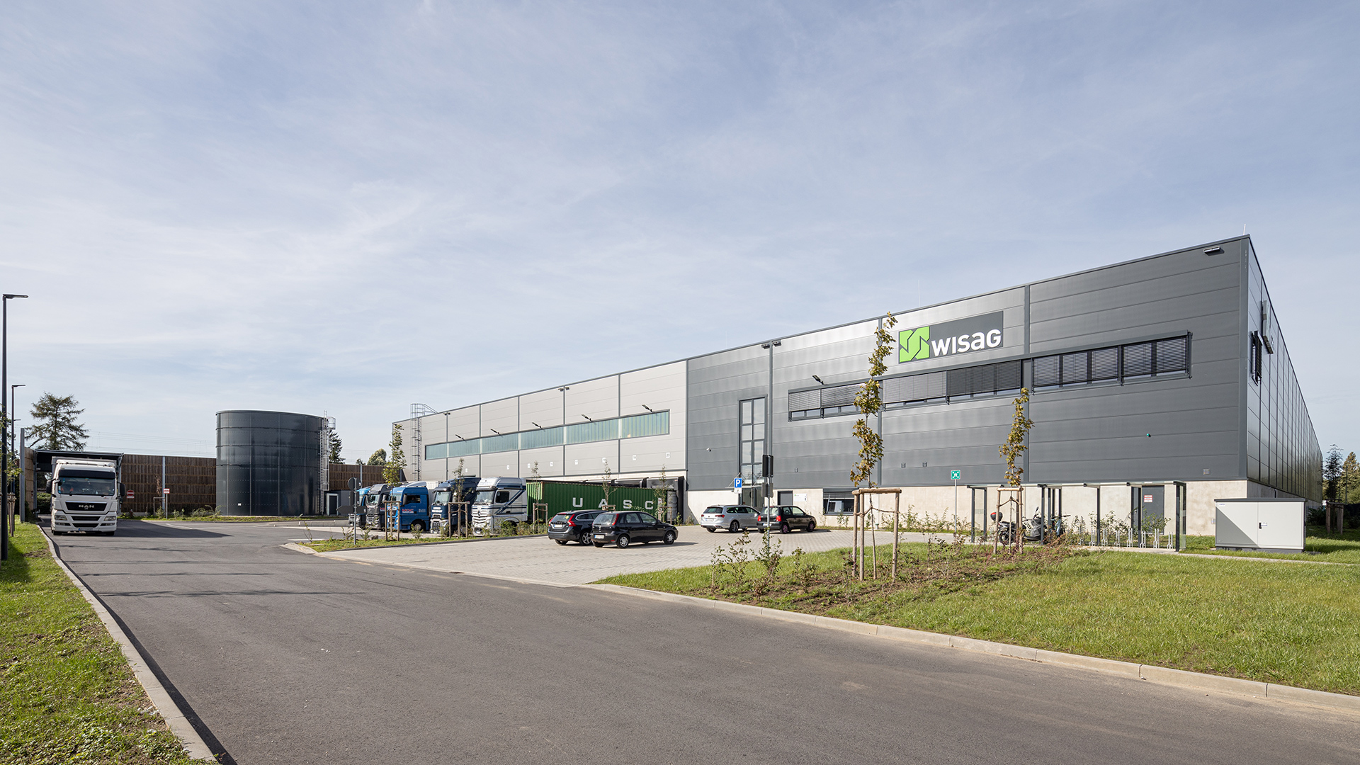 Foto des Wisag-Gebäudes mit Parkplatz, Lkw und grasbewachsener Fläche im Vordergrund unter blauem Himmel.