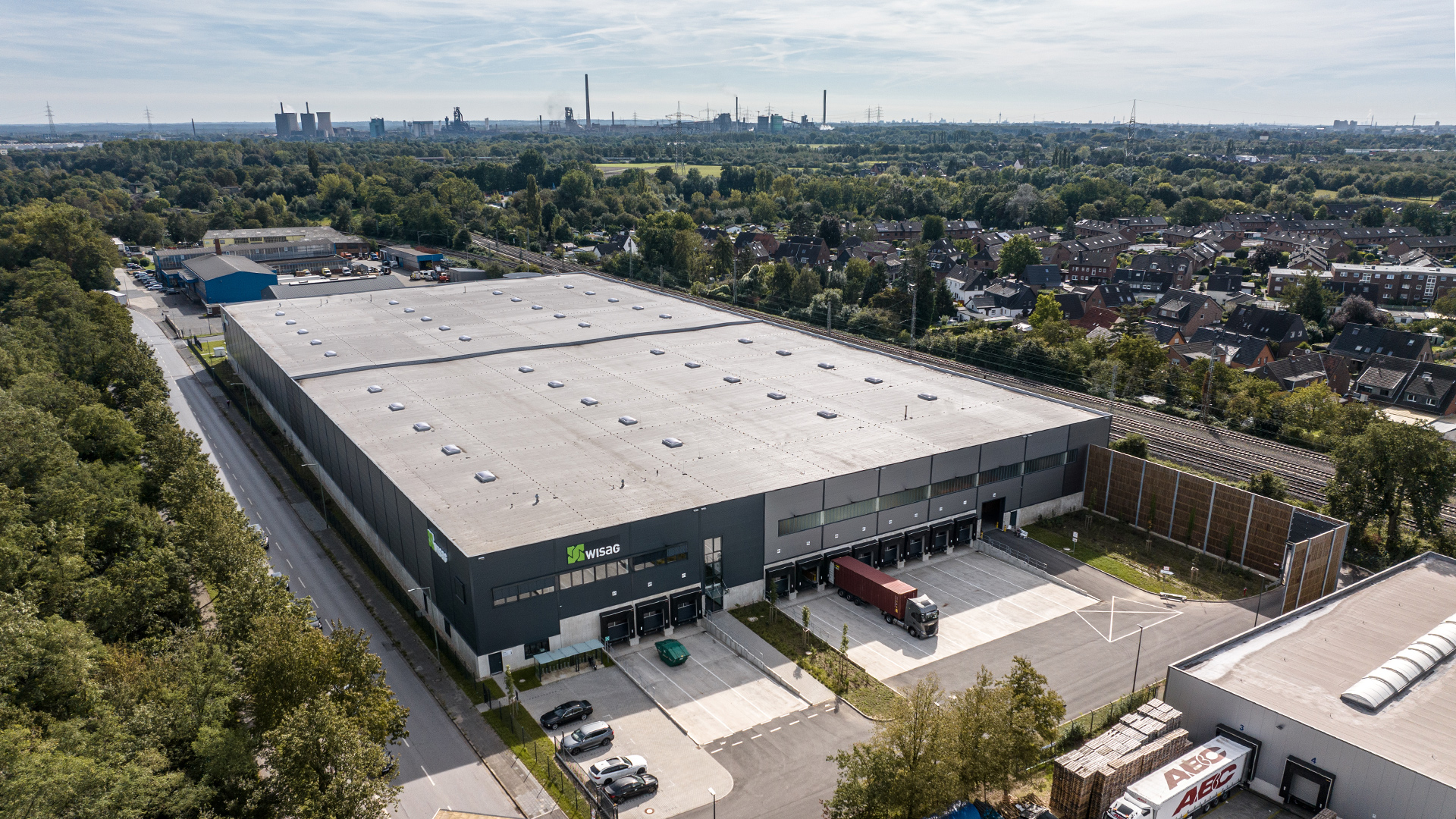 Luftaufnahme eines großen Lagerhauses mit Lkw-Entladung, umgeben von Bäumen und Wohngebieten.