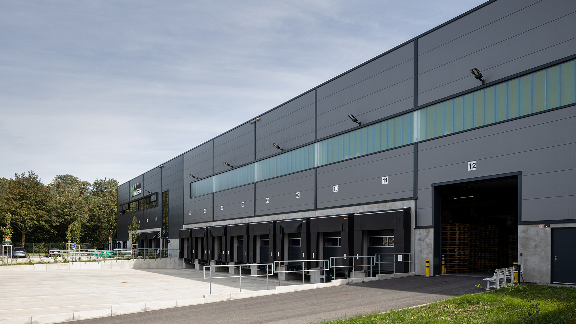 Moderne Lagerhalle mit Lkw-Rampen, großen Fenstern und grünem Umfeld unter blauen Himmel.