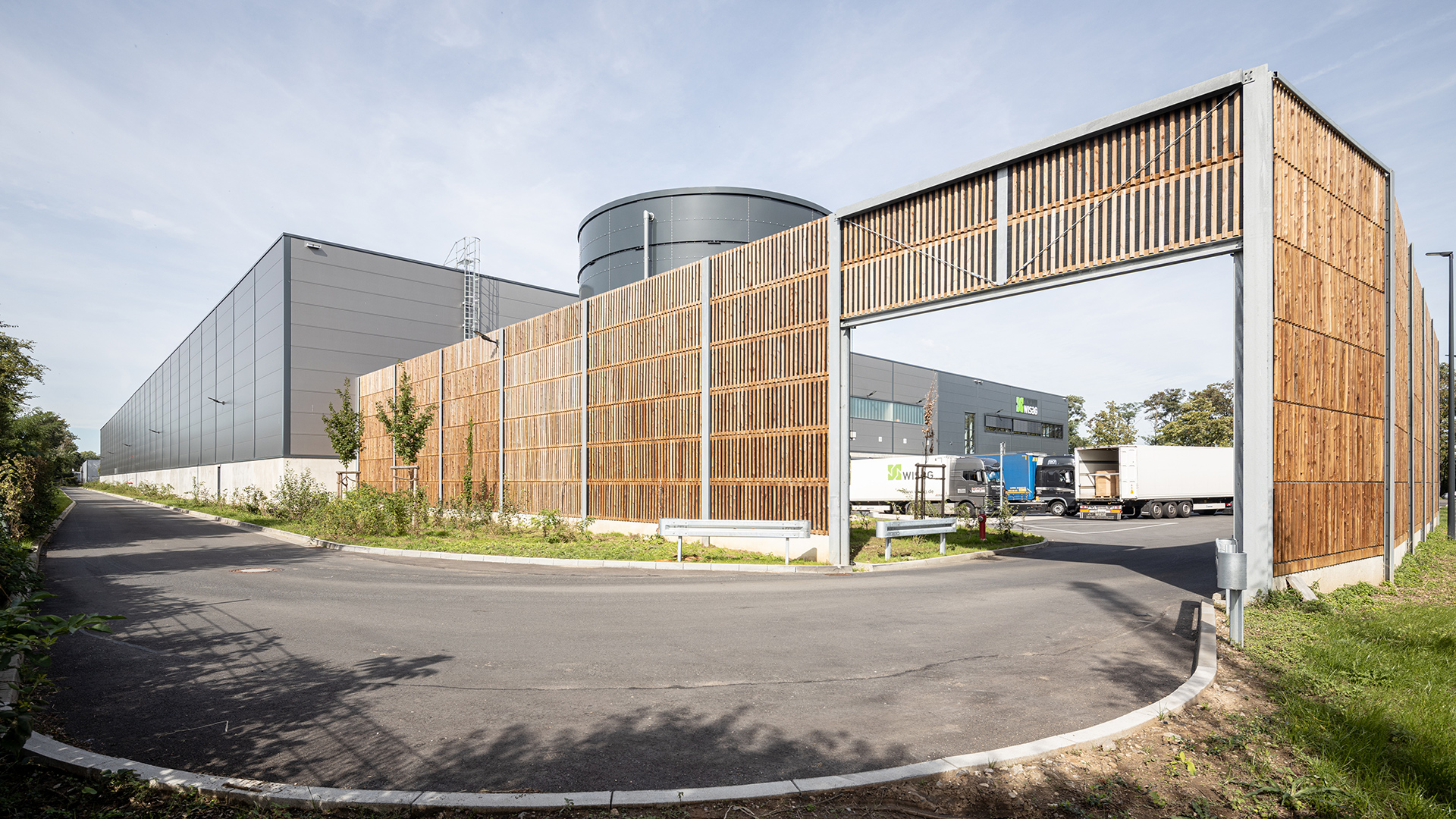 Moderne Lagergebäude mit Holz- und Metallfassade, umgeben von grünen Bereichen und einer Straße.