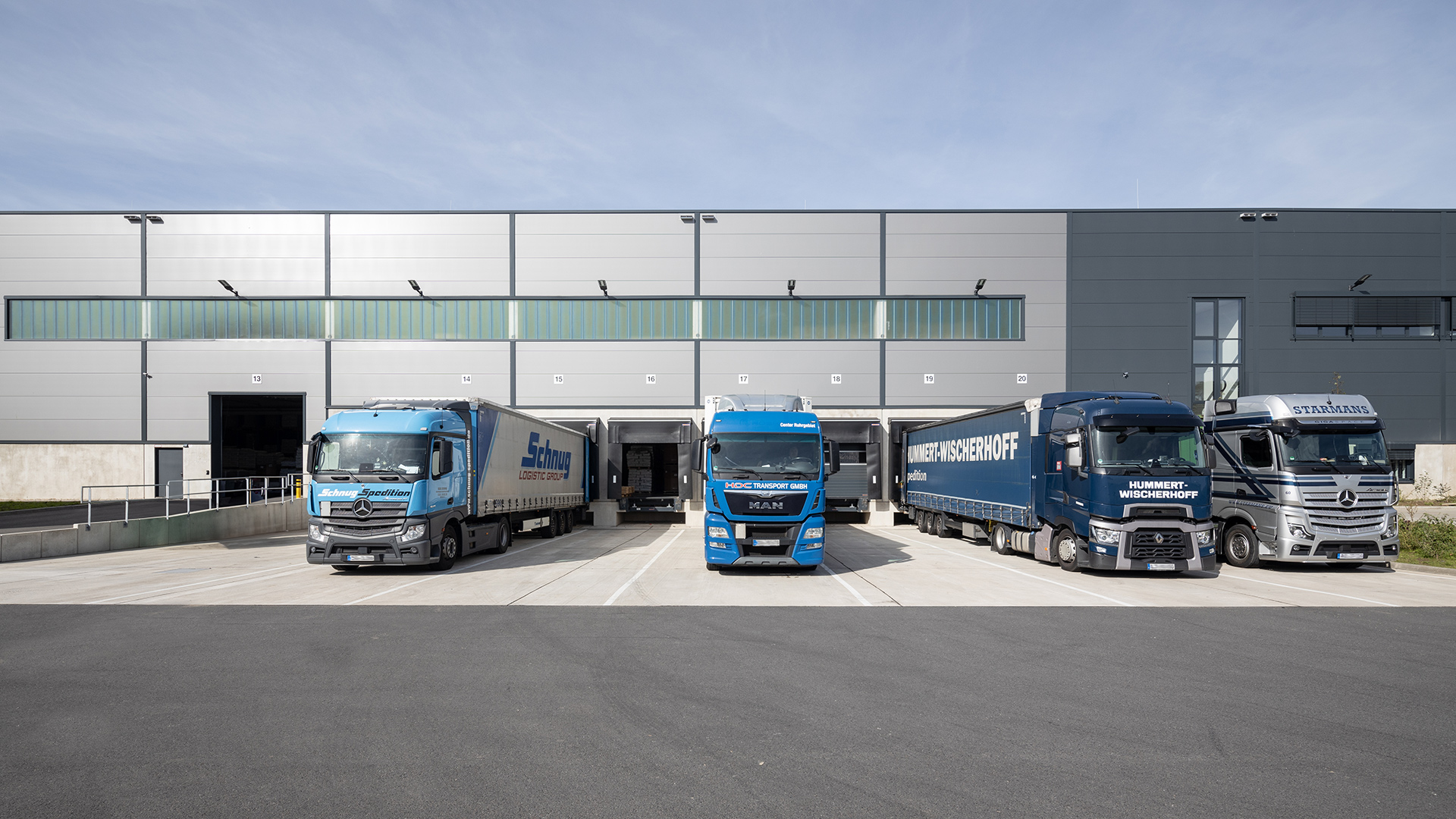 Drei Lastwagen vor einem modernen Lagergebäude, bereit zum Be- und Entladen, bei klarem Himmel.