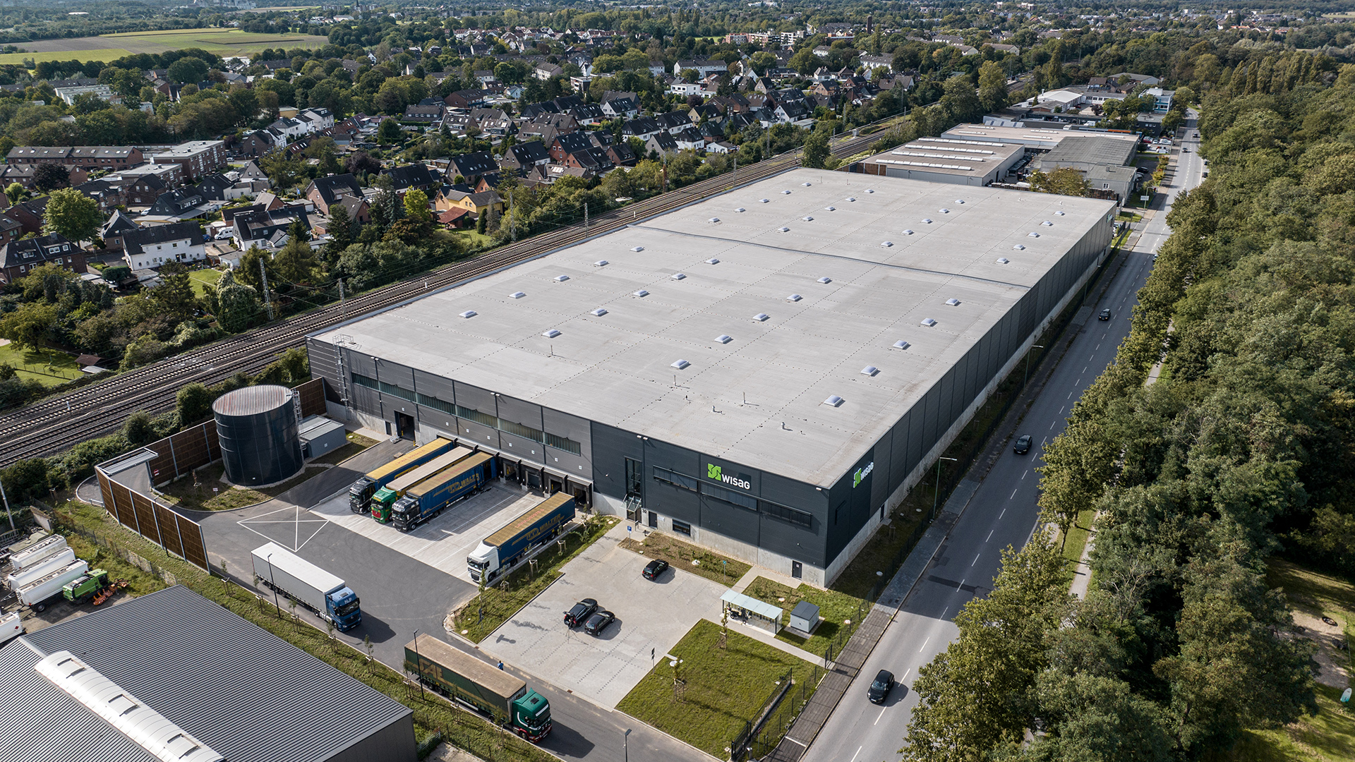Luftaufnahme eines großen Lagerhauses mit Lastwagen und umliegenden Wohngebieten, nahe einer Straße und Eisenbahngleisen.