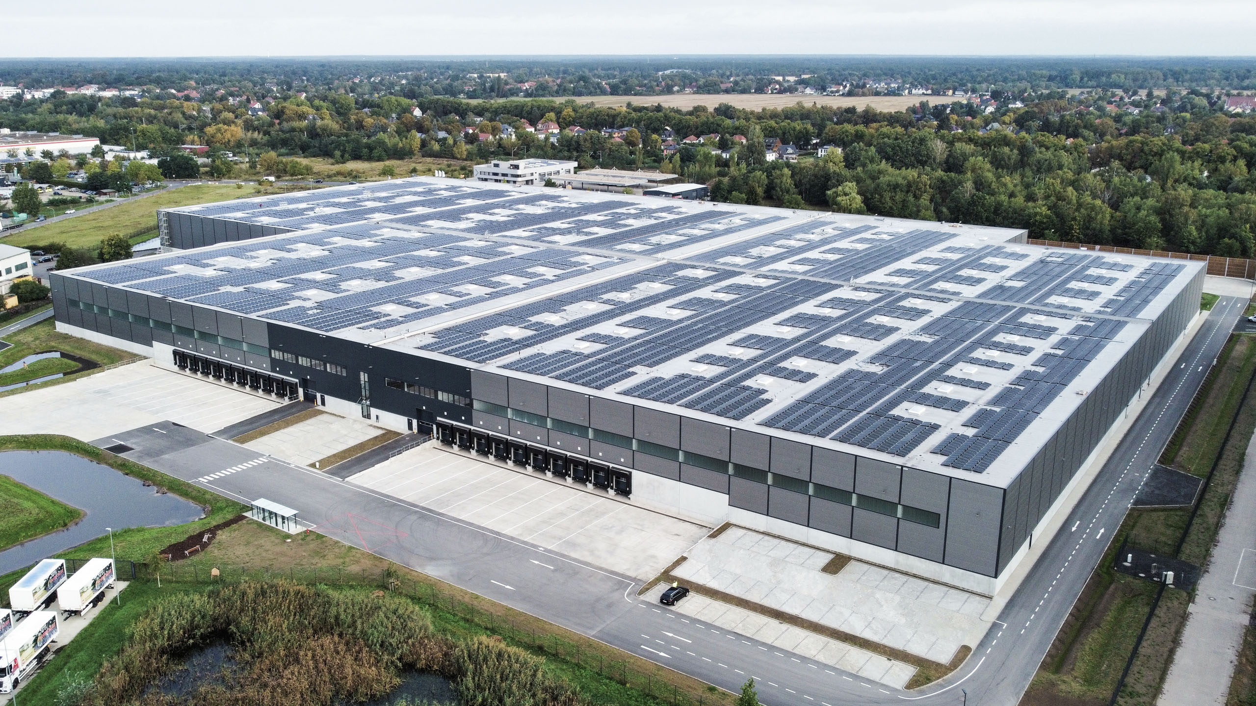 Industriegebäude mit Solarpanelen auf dem Dach, umgeben von grünen Flächen und einem Parkplatz.