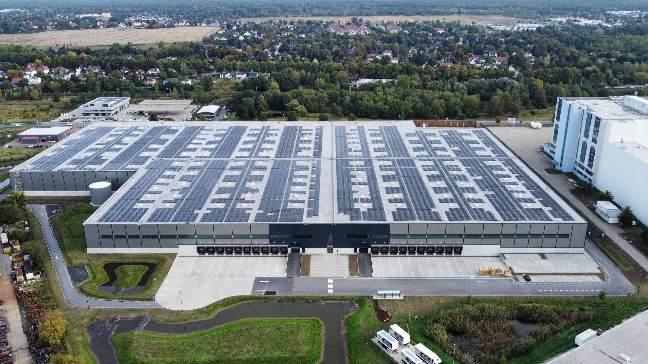 Luftaufnahme eines großen Lagergebäudes mit Solarpanelen, umgeben von Grünflächen und Wohngebieten.