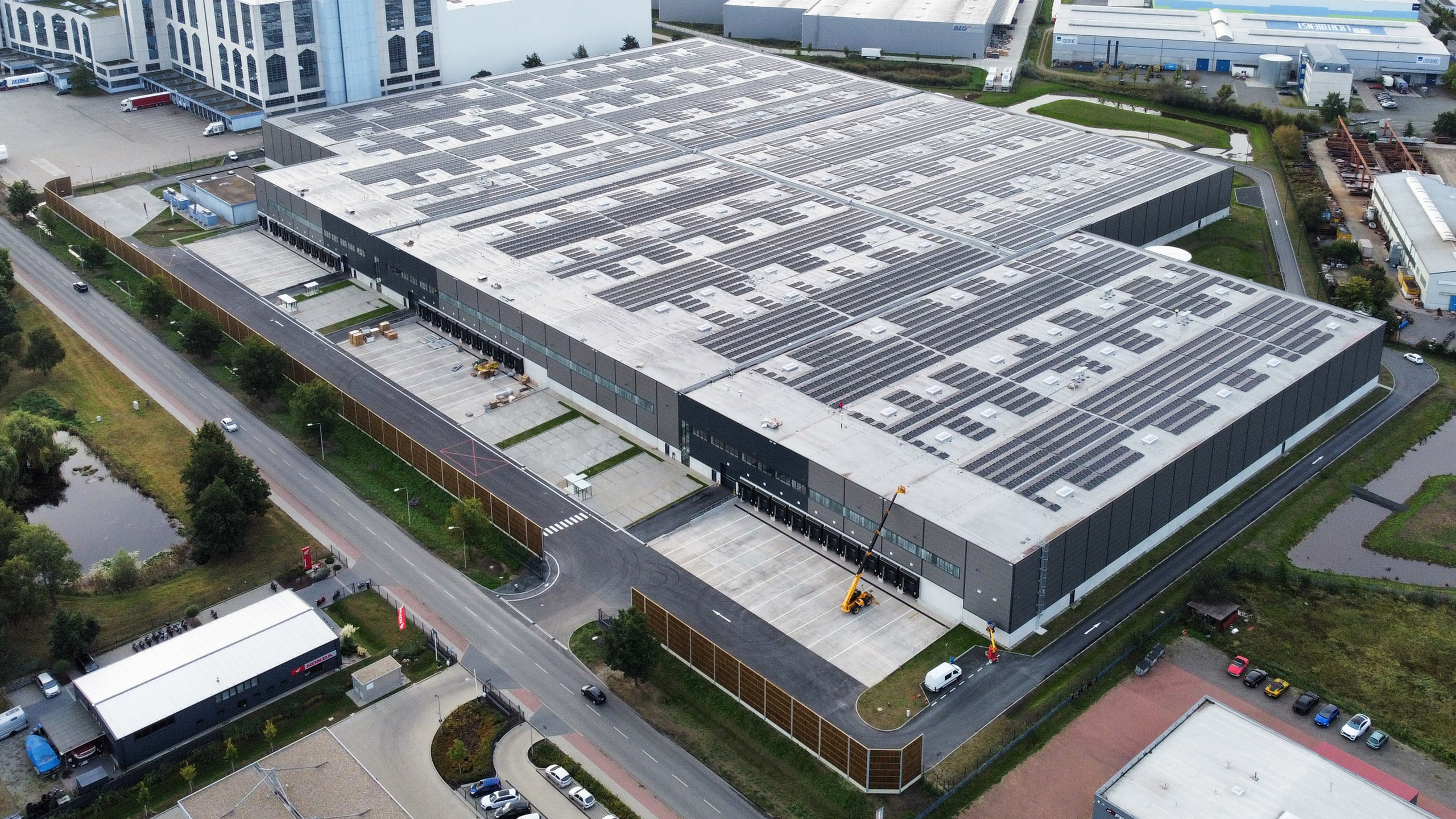 Großes Lagergebäude mit Solarpanelen auf dem Dach, umgeben von Straßen und Landschaft. Reifenspeicher und Lkw-Anlieferung sichtbar.