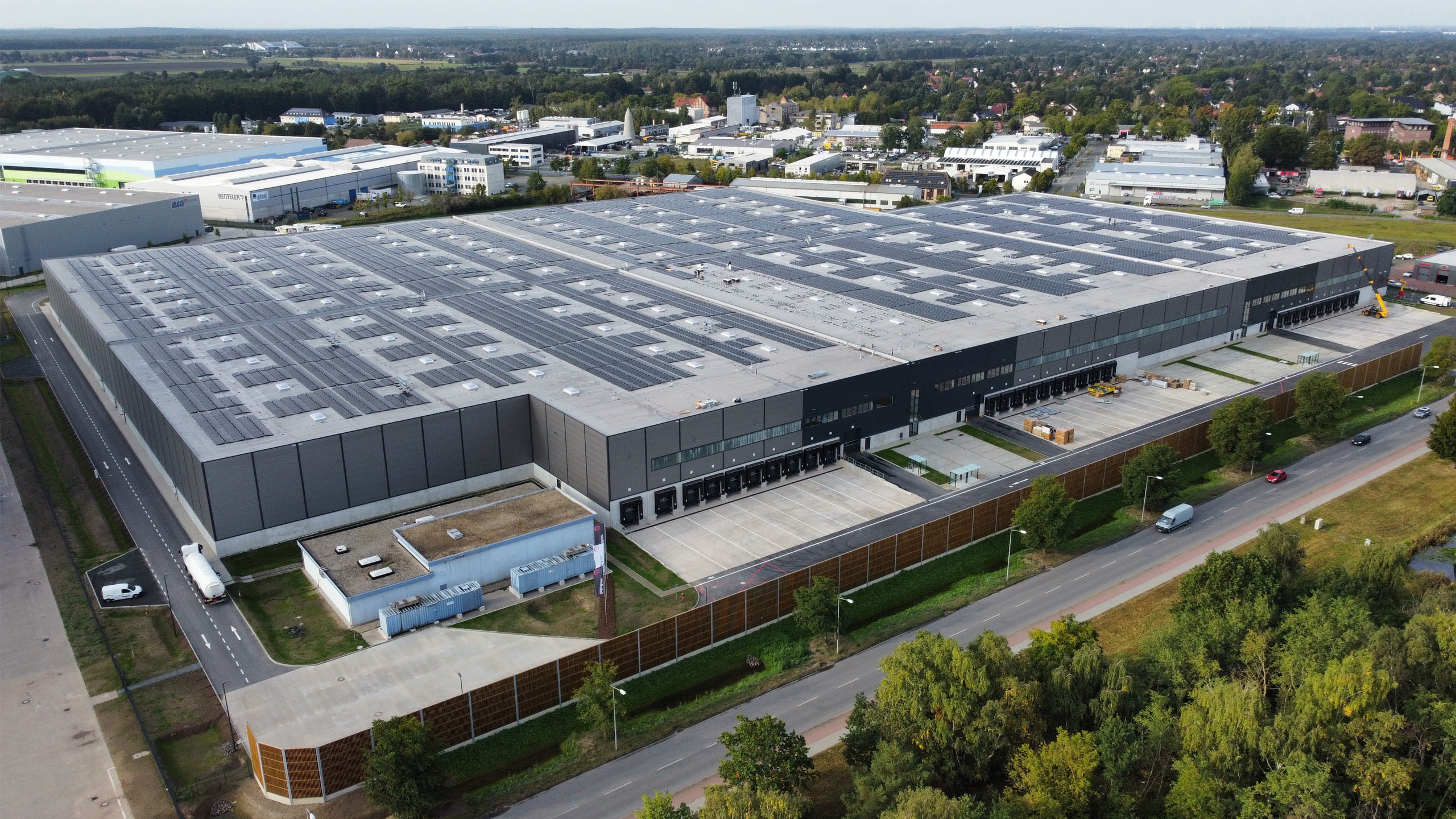 Modernes Lagerhaus mit Solaranlagen auf dem Dach, umgeben von Straßen und Bäumen, in einer städtischen Umgebung.