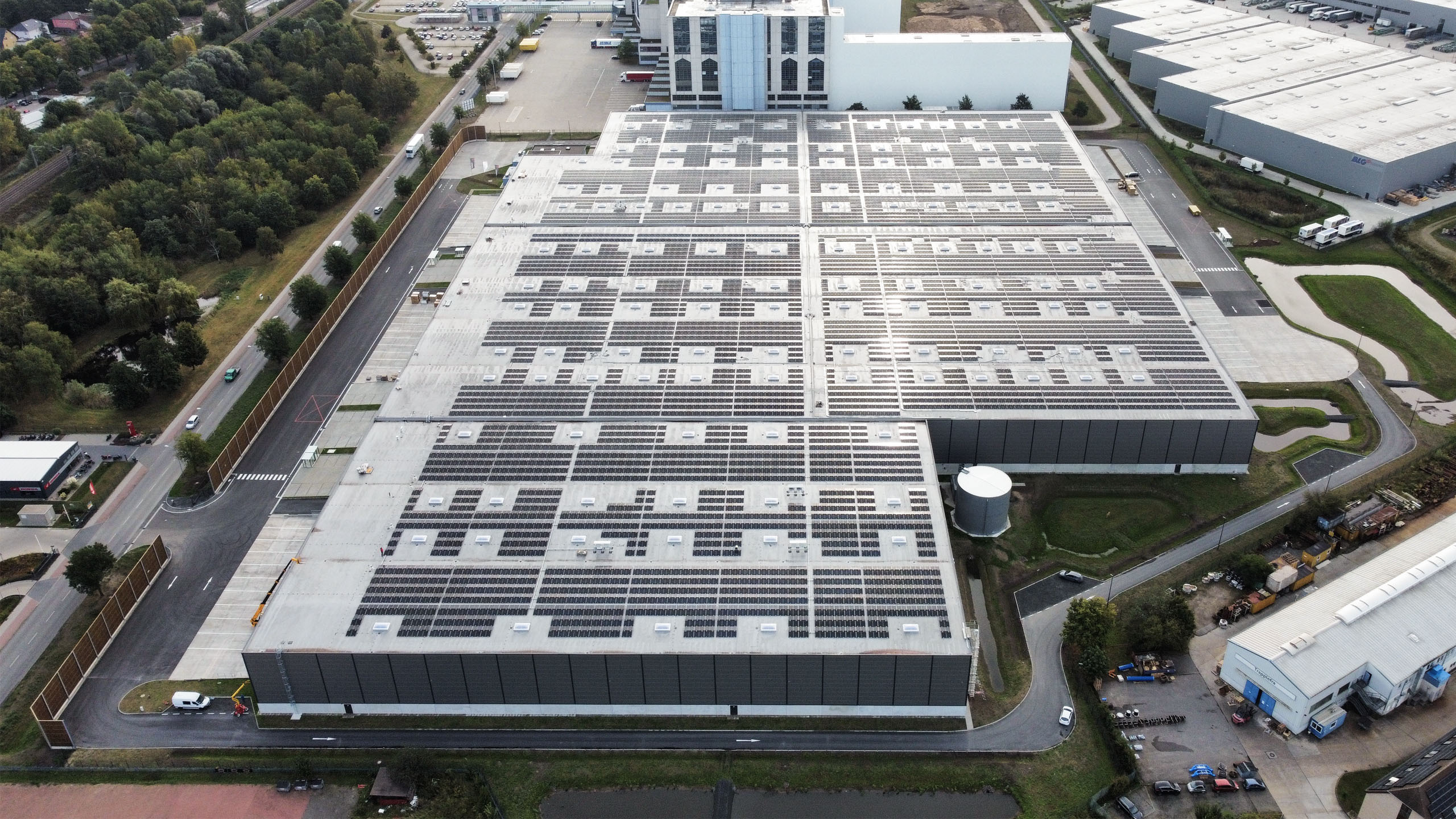 Luftaufnahme eines Gebäudes mit Solaranlagen auf dem Dach, umgeben von Straßen und Grünflächen.