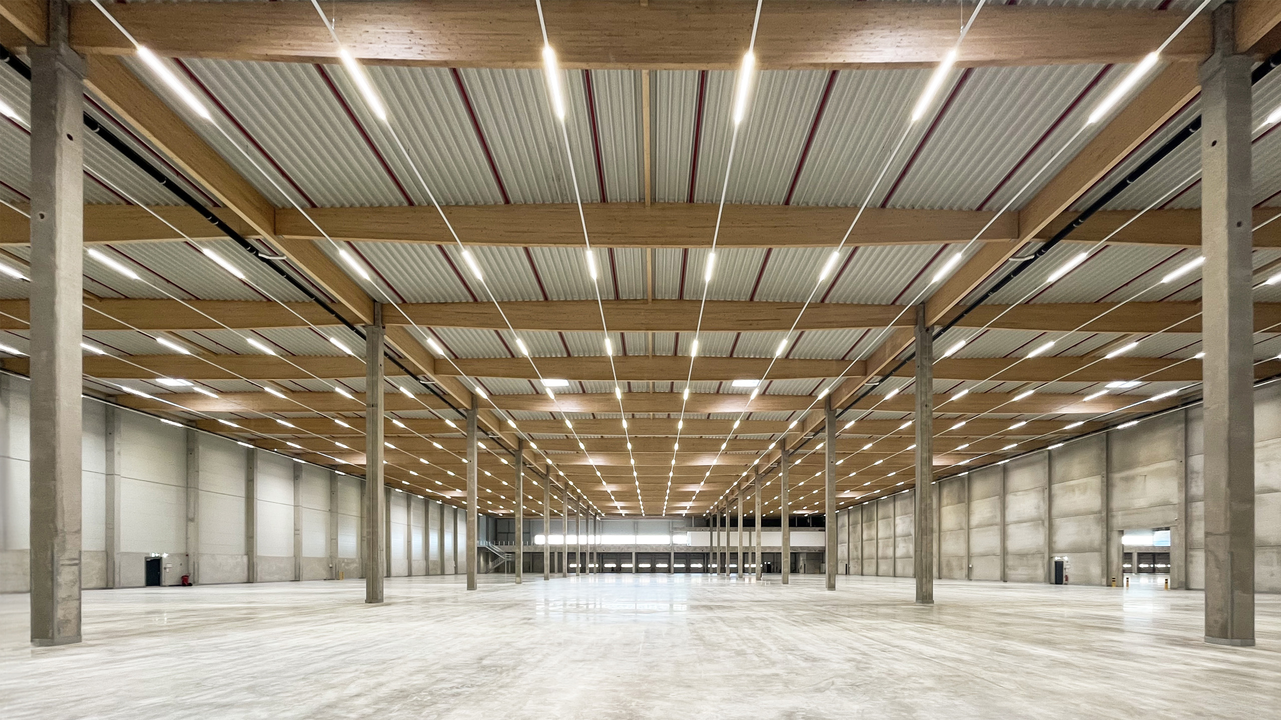 Ein weites, leeres Lager mit Holzbalken und Deckenbeleuchtung, betonierten Wänden und einem glatten, hellen Boden.