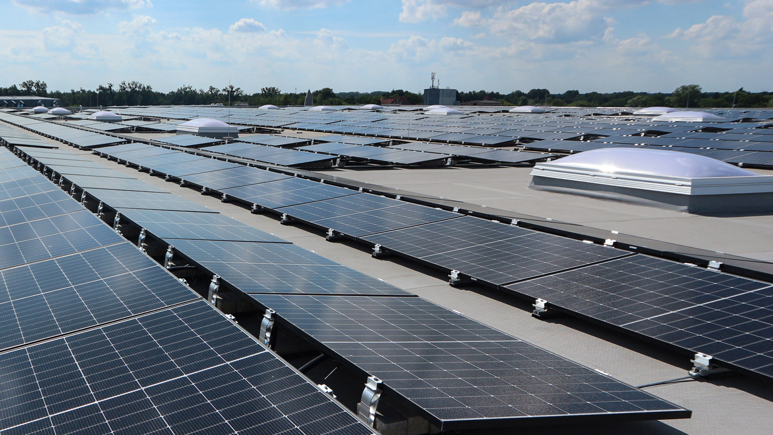 Solaranlage auf einem Dach, mit zahlreichen Solarzellen in einem blauen Himmel und einigen Lichtkuppeln im Hintergrund.