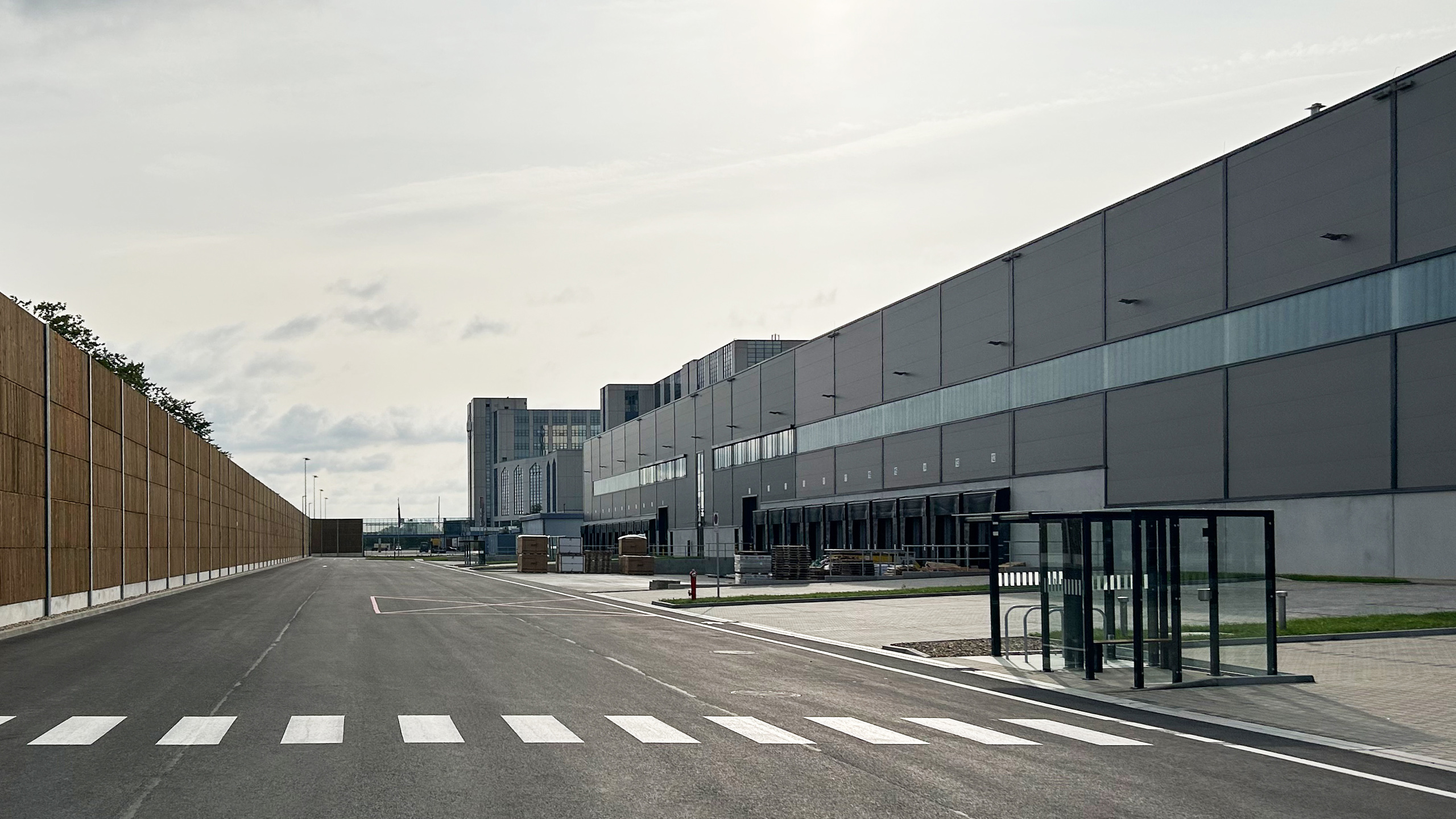 Blick auf eine moderne Lagerhalle mit angrenzender verkehrsberuhigter Straße und Bushaltestelle. Wolkenbedeckter Himmel im Hintergrund.
