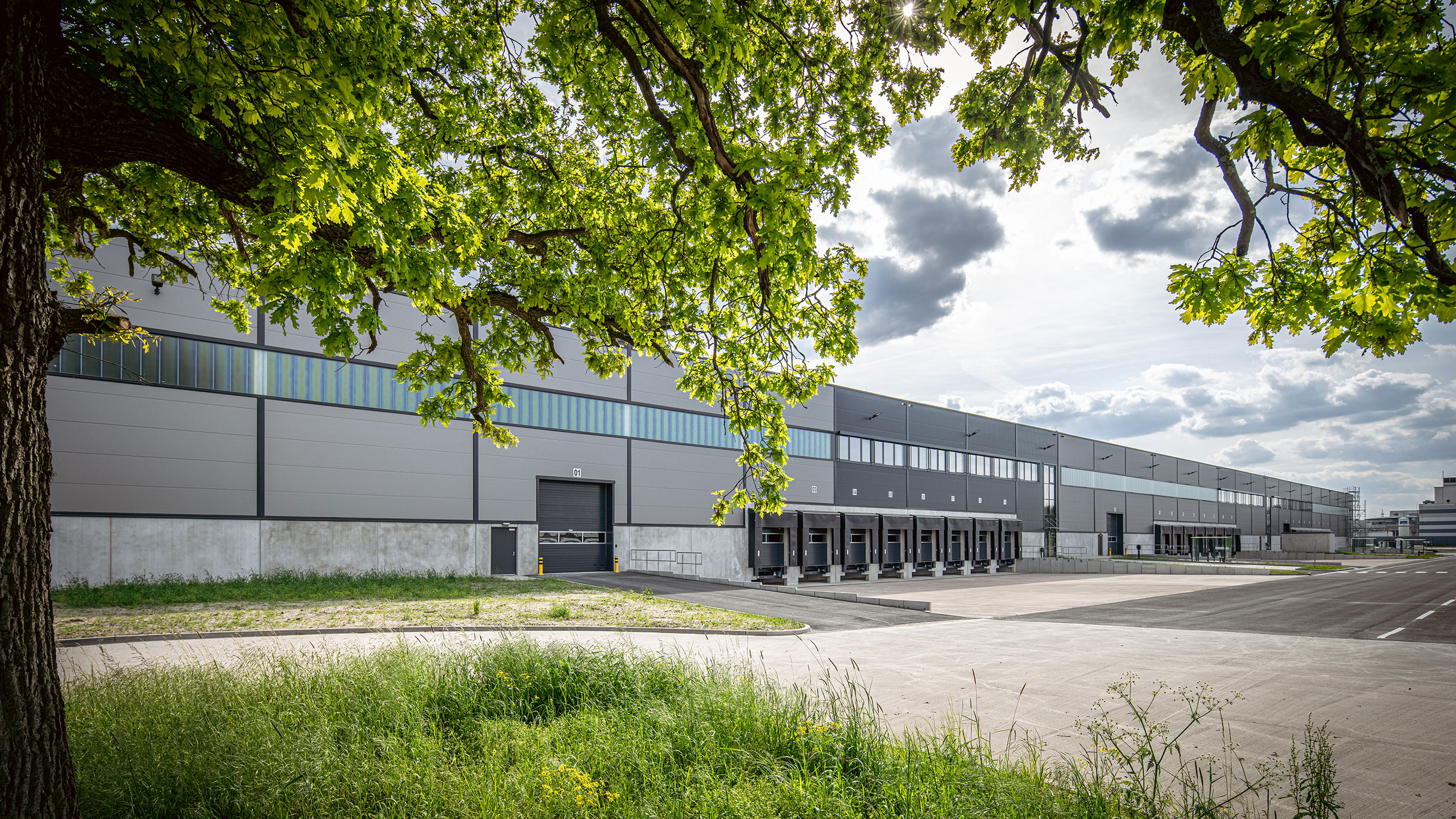 Ein moderner Lagerhauskomplex mit Verladetoren, umgeben von Bäumen und Gras unter einem bewölkten Himmel.
