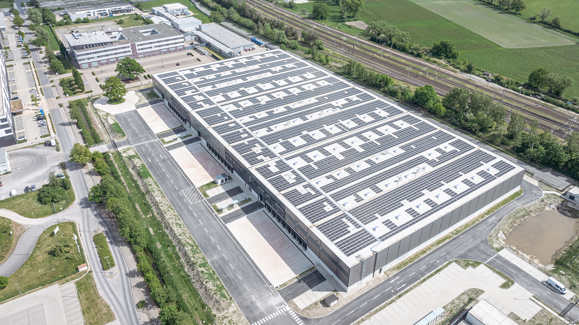 Luftaufnahme eines modernen Gebäudes mit Solarpanels, umgeben von Straßen und Grünflächen.